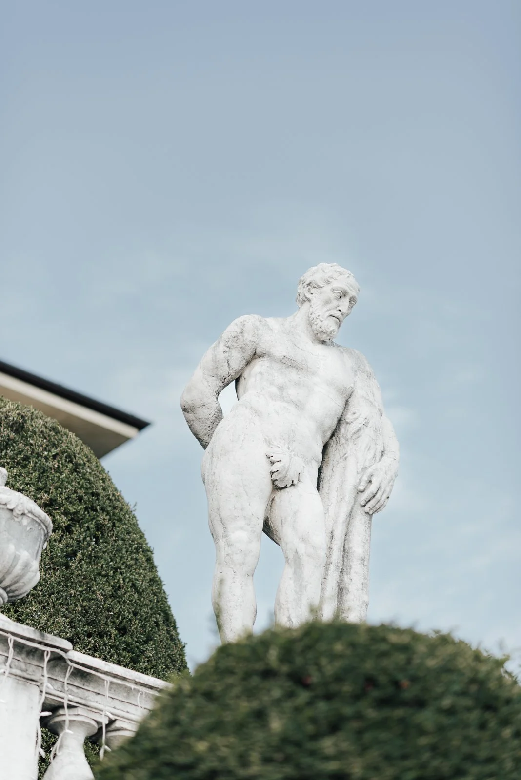 Eine weiße Skulptur eines männlichen nackten Körpers mit Bart, die auf einem Podest steht, umgeben von grünen Büschen unter einem blauen Himmel.