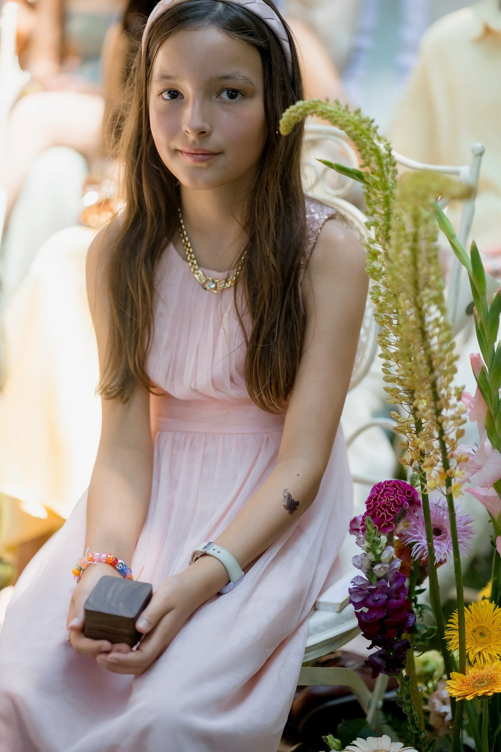 Ein junges Mädchen in einem rosa Kleid sitzt auf einer Bank mit bunten Blumen im Hintergrund. Sie trägt eine goldene Halskette, eine Armbanduhr und einen bunten Armreif. Sie hält ein Smartphone in den Händen und lächelt leicht. Das Mädchen hat lange 