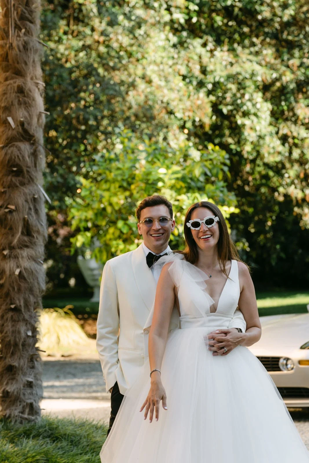Ein Hochzeitsfoto eines glücklichen Paares im Freien mit grünen Bäumen im Hintergrund. Die Braut trägt ein weißes Hochzeitskleid und Sonnenbrille, während der Bräutigam einen weißen Anzug mit Fliege trägt.