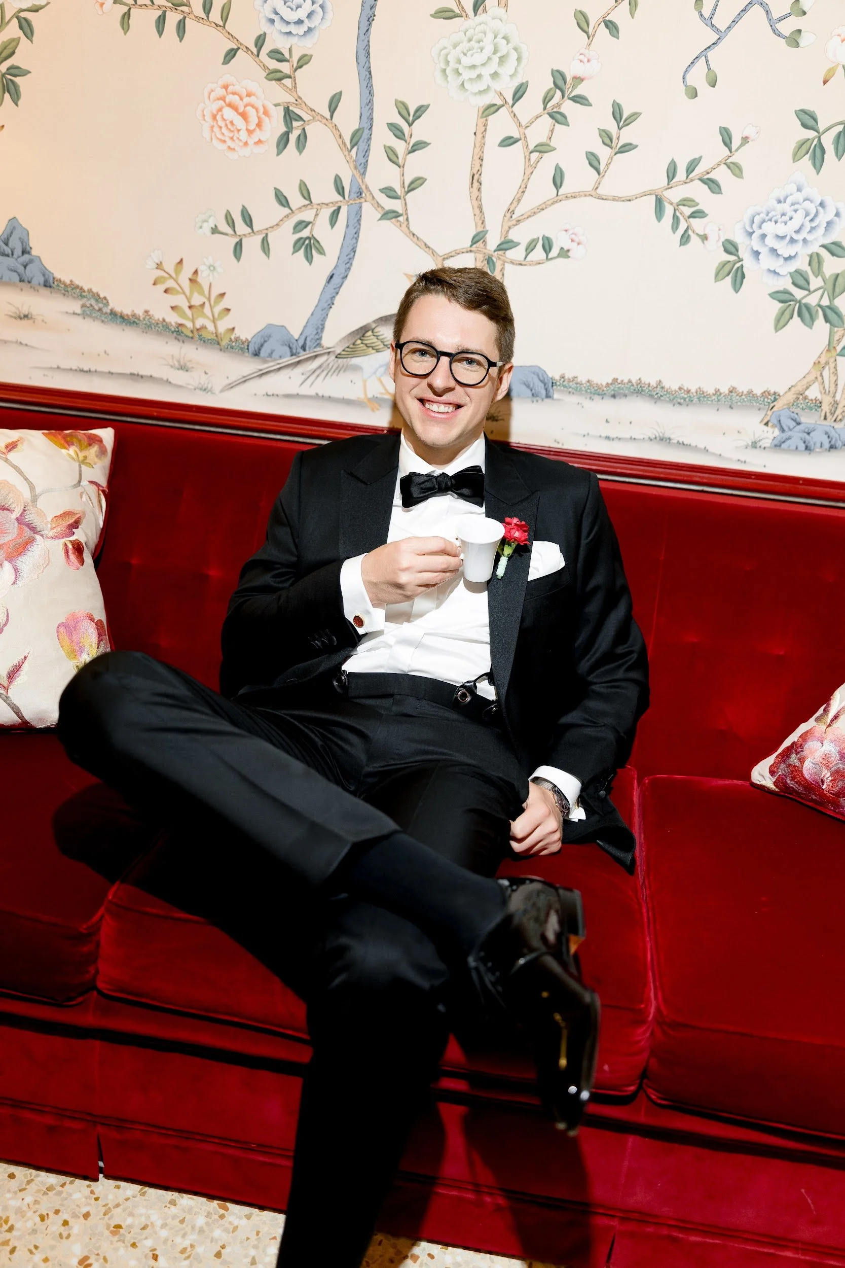 Junger Mann in Smoking mit Fliege, sitzt auf rotem Sofa, hält eine kleine weiße Tasse und lächelt. Hinter ihm eine Wand mit bemalter Landschaft, Bäumen und Blumen, sowie ein Kissen mit Blumenmuster.