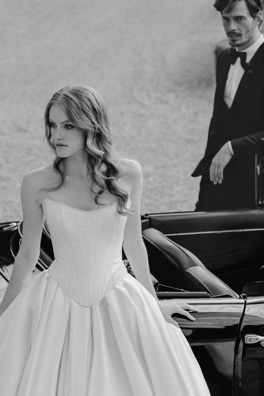 Schwarz-weiß-Fotografie eines Paares, eine Frau in einem eleganten Ballkleid, die neben einem Cabrios sitzt, und ein Mann in einem schwarzen Anzug mit Fliege, der neben dem Auto steht.
