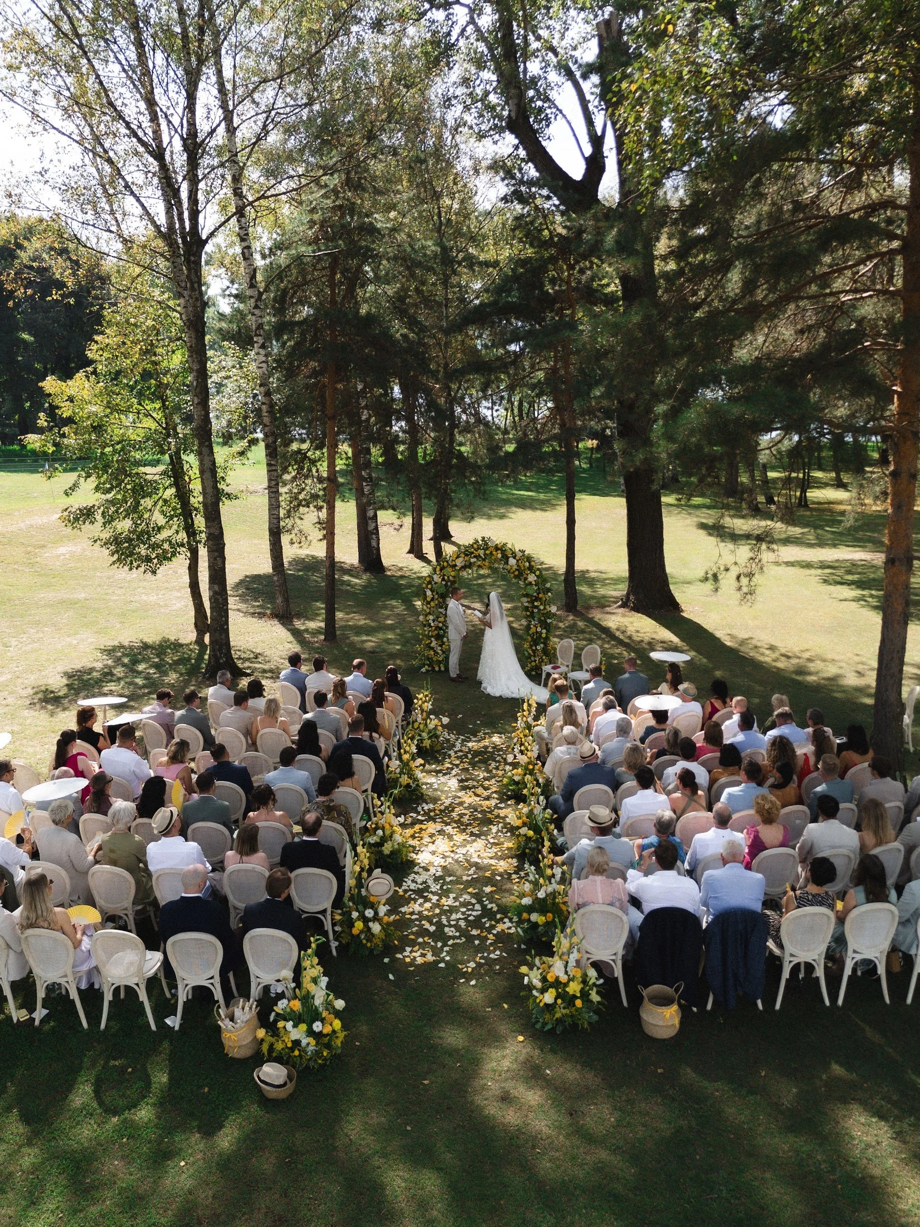 Ein Garten bei Sonnenlicht, in dem eine Hochzeit im Freien stattfindet. Ein Brautpaar steht unter einem Blumenbogen, umgeben von Gästen, die in Stühlen sitzen.
