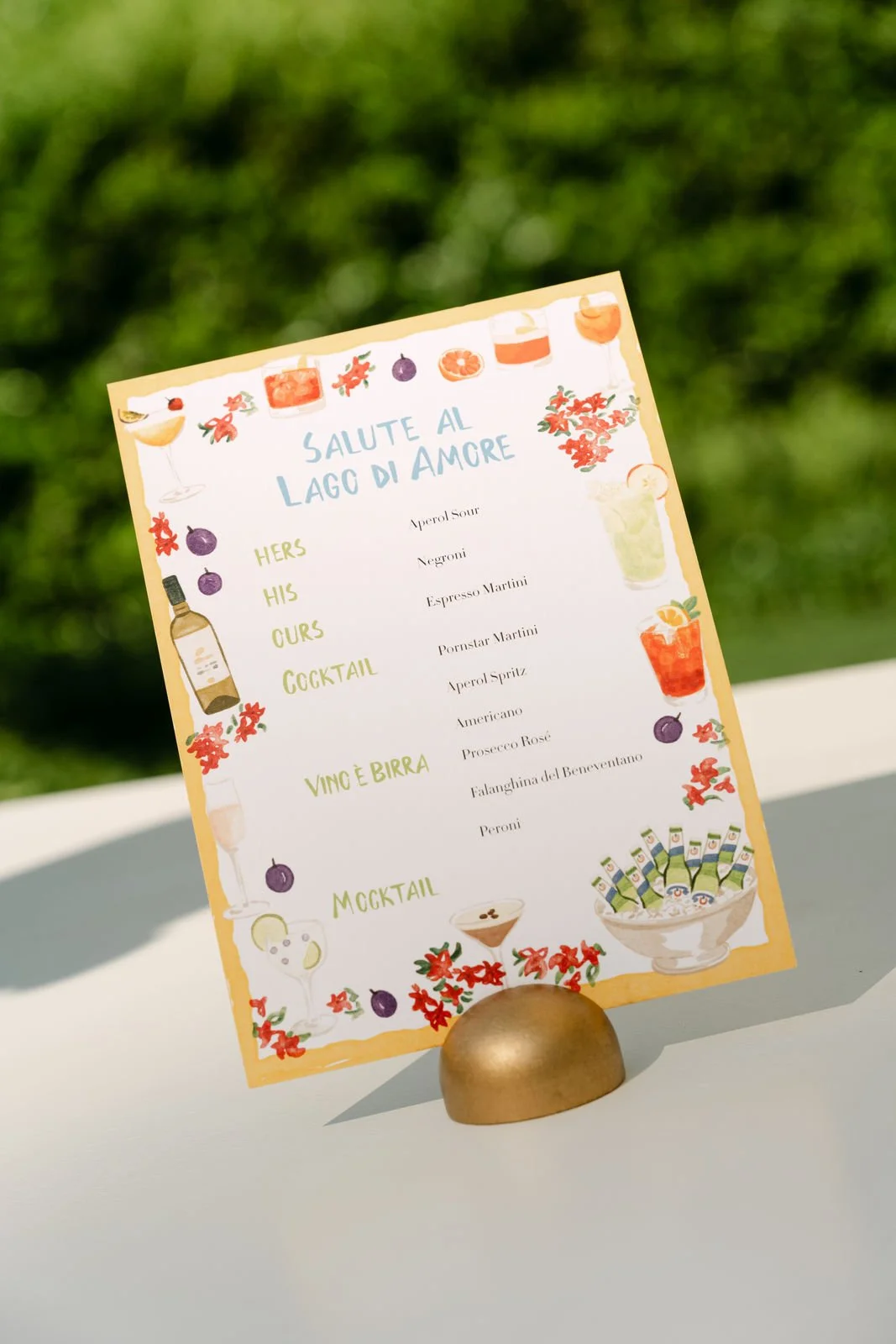 Getränkekarte mit italienischen Cocktails und Bieren, dekoriert mit bunten Illustrationen von Getränken und Früchten, auf einem weißen Tisch im Freien.