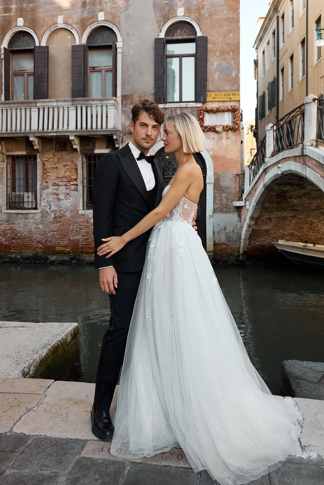 Ein Paar in Hochzeitskleidung steht an einem Kanal in Venedig, Italien. Der Mann trägt einen schwarzen Anzug mit Fliege, die Frau trägt ein weißes Brautkleid mit Tüllrock.