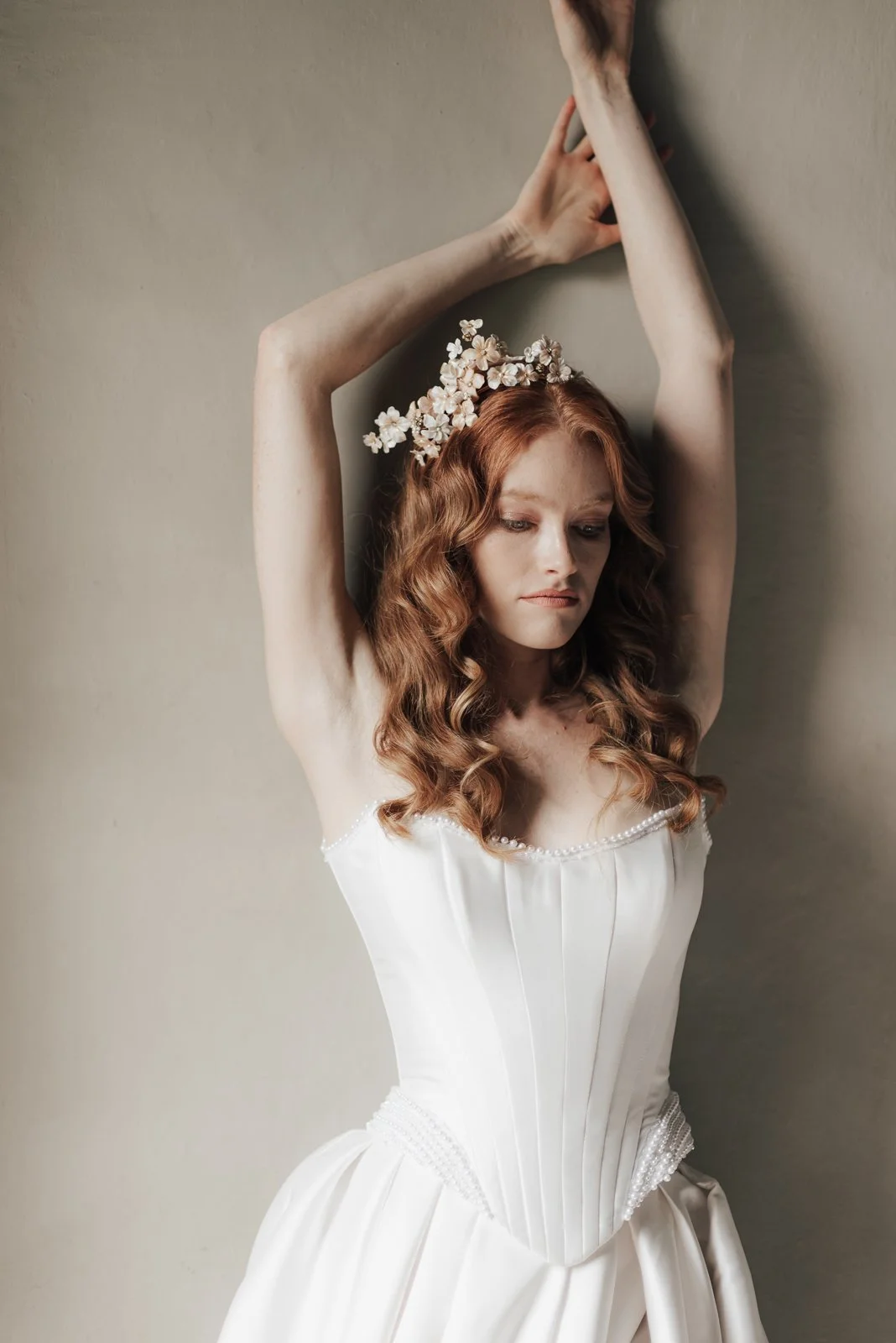 Frau in Hochzeitskleid mit Blumenkranz im Haar, die an einer Wand lehnt.
