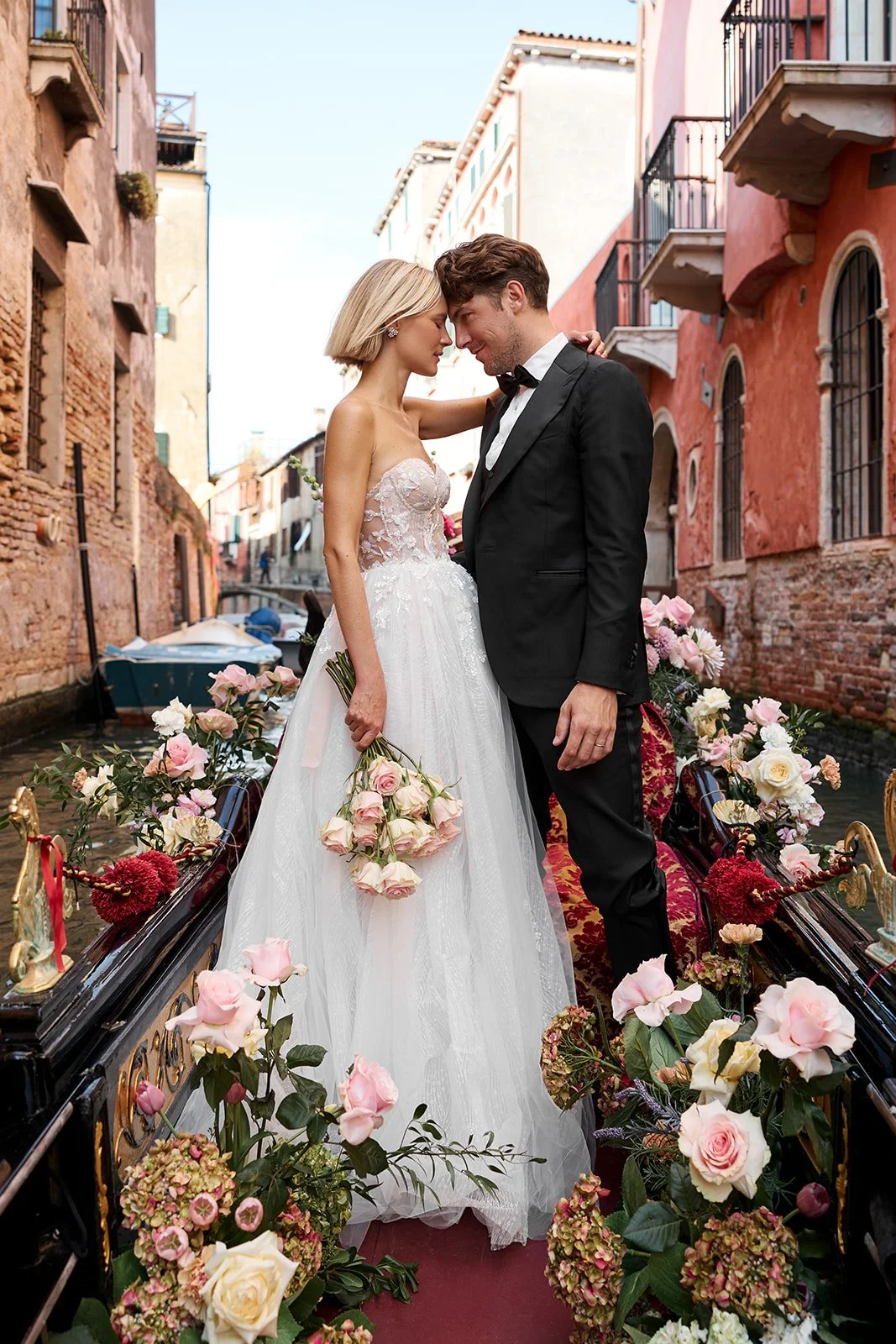 Ein Hochzeitsfoto eines Paares in formeller Kleidung, die auf einer Blumenverzierten Kutsche in einer Kanalszene in Venedig stehen, umgeben von bunten Häusern und Wasser.