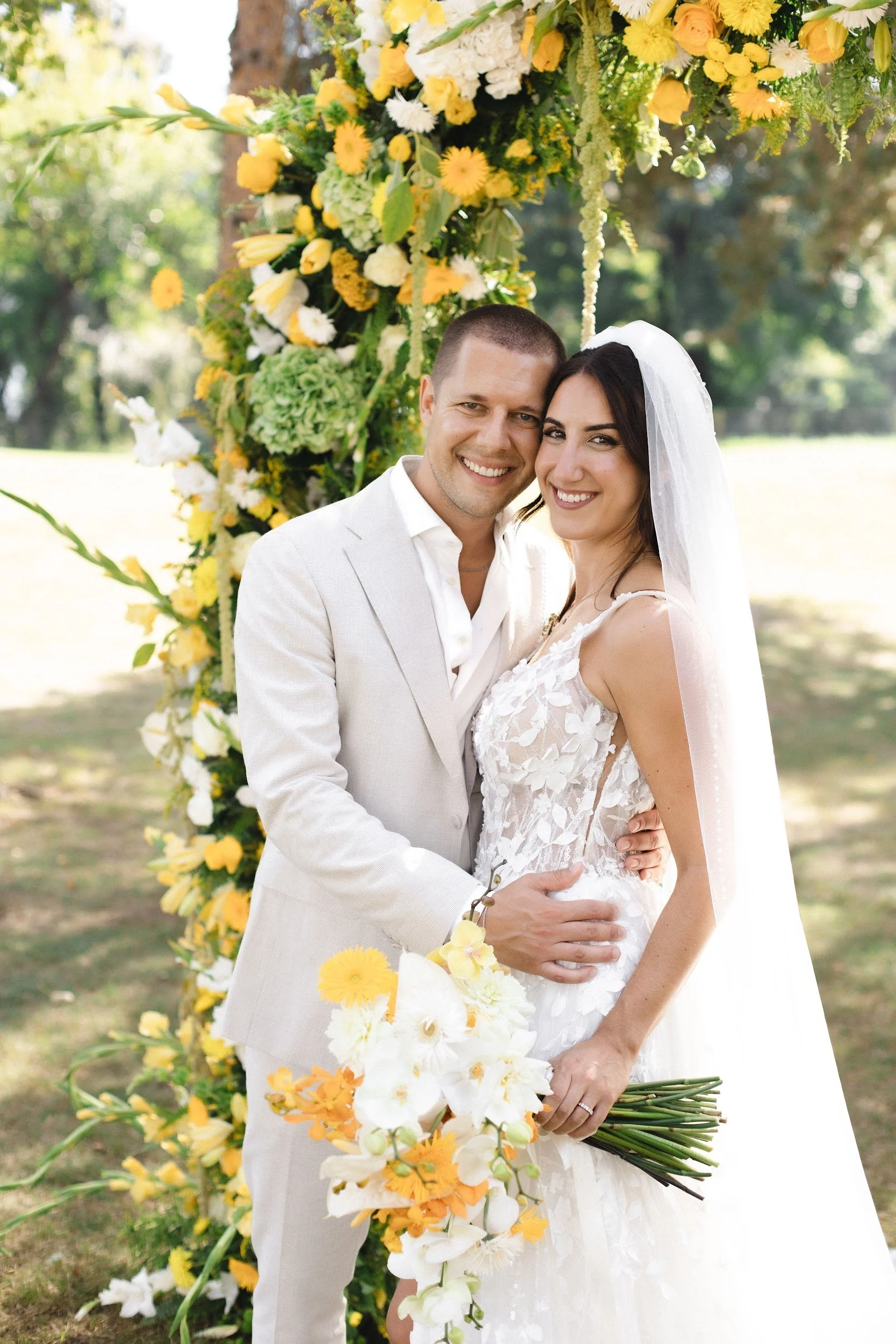 Ein glückliches Brautpaar bei einer Hochzeit im Freien, umgeben von einer Blumenpracht aus weißen, gelben und orangefarbenen Blumen.