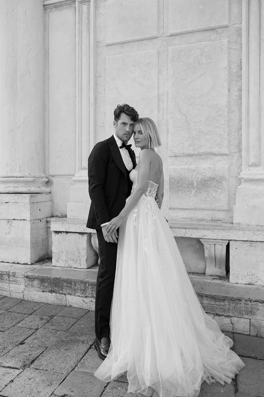 Ein Mann und eine Frau in formeller Kleidung, vermutlich für eine Verlobung oder Hochzeit, stehen vor einer alten Steinmauer. Der Mann trägt einen schwarzen Anzug mit Fliege, die Frau trägt ein langes, elegantes weißes Kleid.