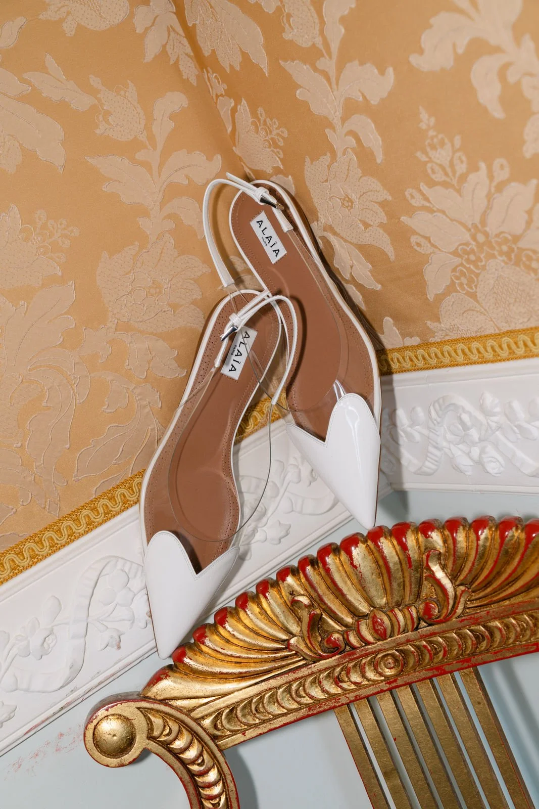 Ein Paar weiße High Heels mit transparentem Bereich und Schnalle auf einem goldfarbenen, floralen Musterhintergrund.