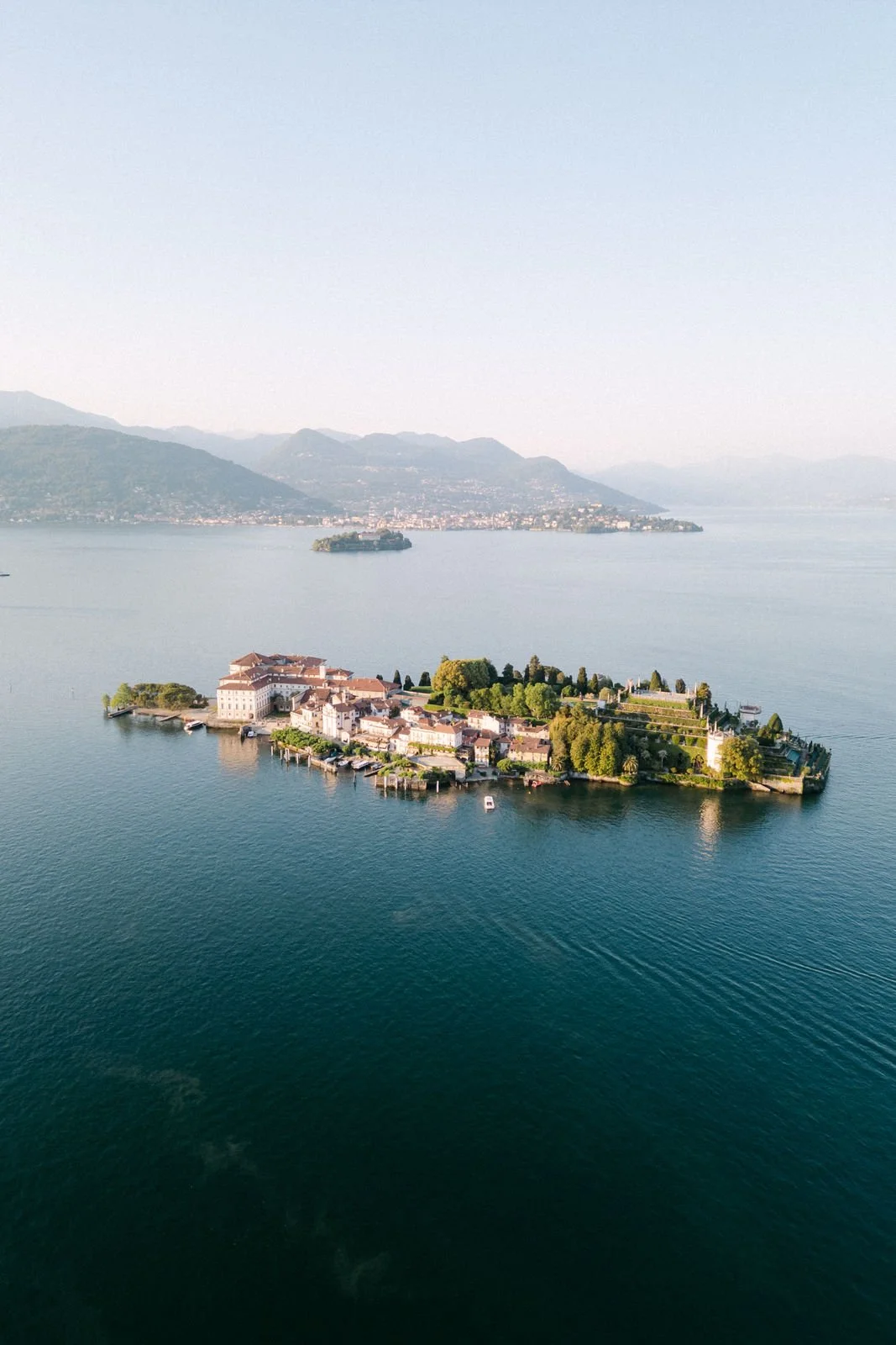 Ein Luftbild zeigt eine kleine Insel mit historischen Gebäuden, umgeben von einem See, mit Bergen im Hintergrund.