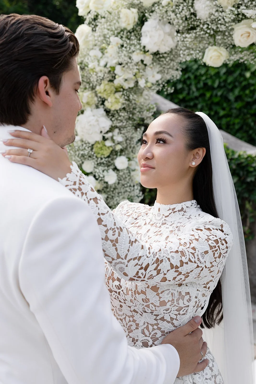 Ein Brautpaar bei ihrer Hochzeit, die Frau trägt ein weißes Spitzenkleid und eine Schleier, der Mann trägt einen weißen Anzug, im Hintergrund sind weiße Rosen und Blumen.