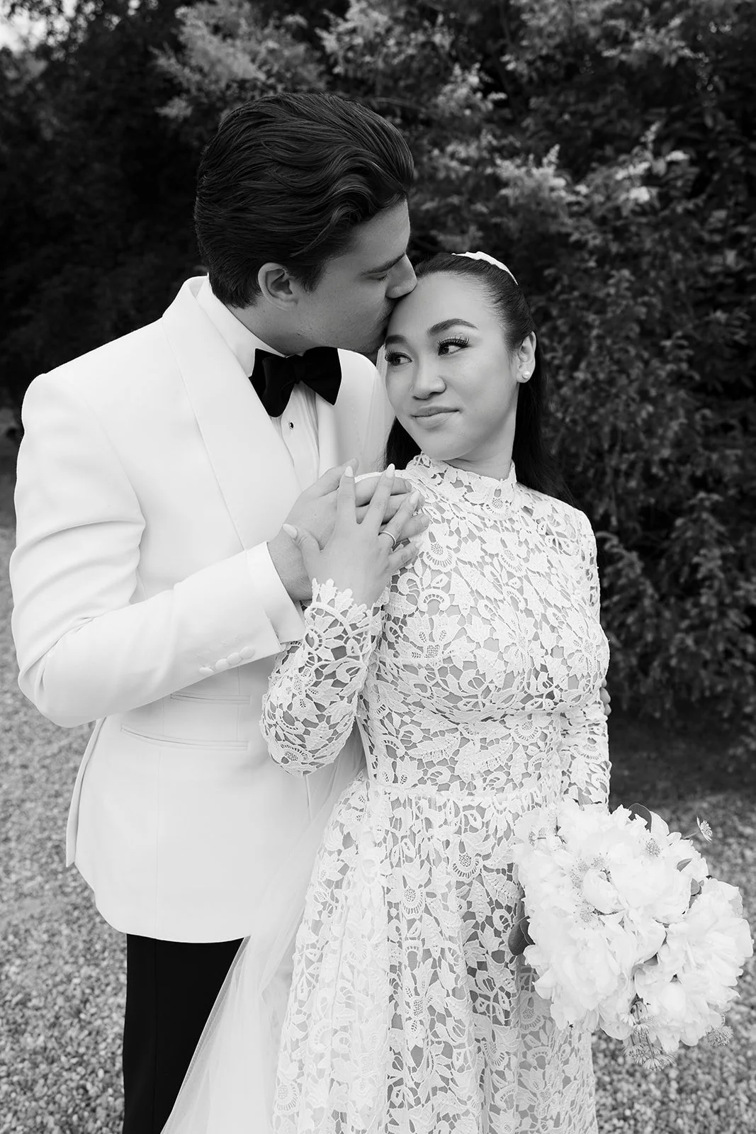 Ein schwarzer und weißer Hochzeitsfoto eines Paares, das sich küsst, während die Braut eine Hochzeitstracht und einen Blumenstrauß hält.
