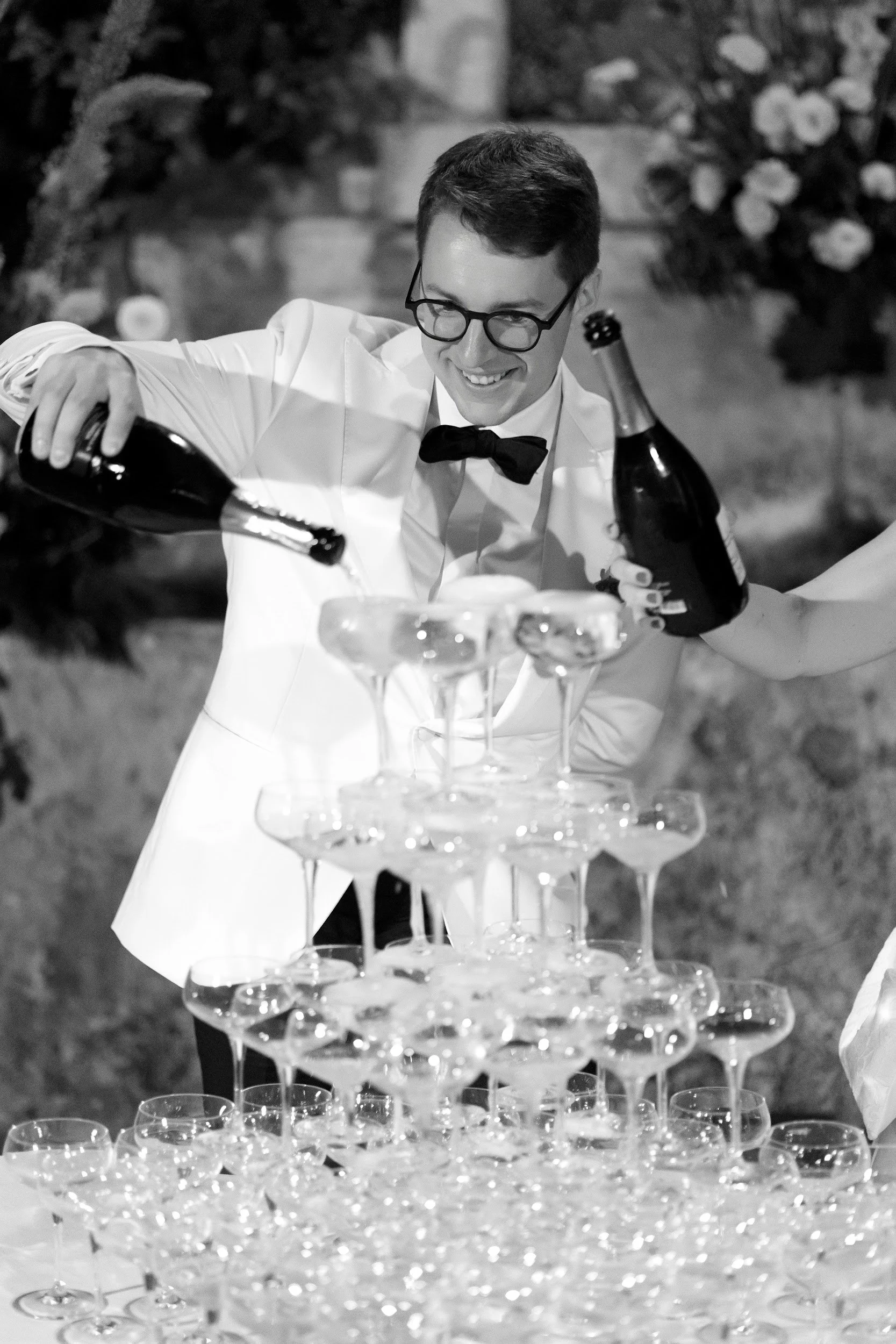 Ein junger Mann in formeller Kleidung, der einen weißen Smoking und eine schwarze Fliege trägt, steht hinter einer Pyramide aus leeren Champagnergläsern und gießt Champagner ein.