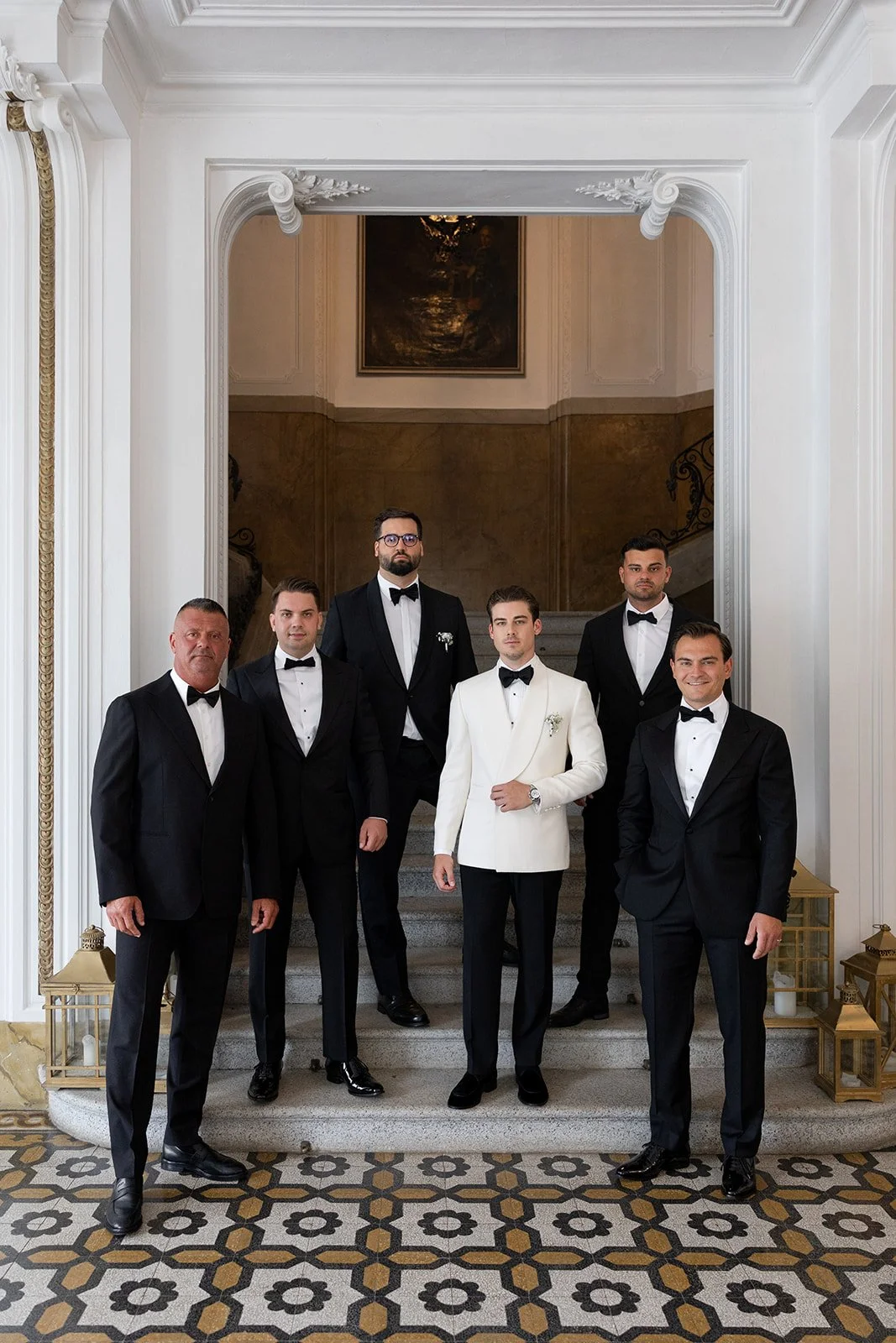 Männer in eleganten Anzügen und Smokings stehen auf einer Treppe in einem prächtigen, barocken Raum mit weißen Verzierungen und Mosaikboden.