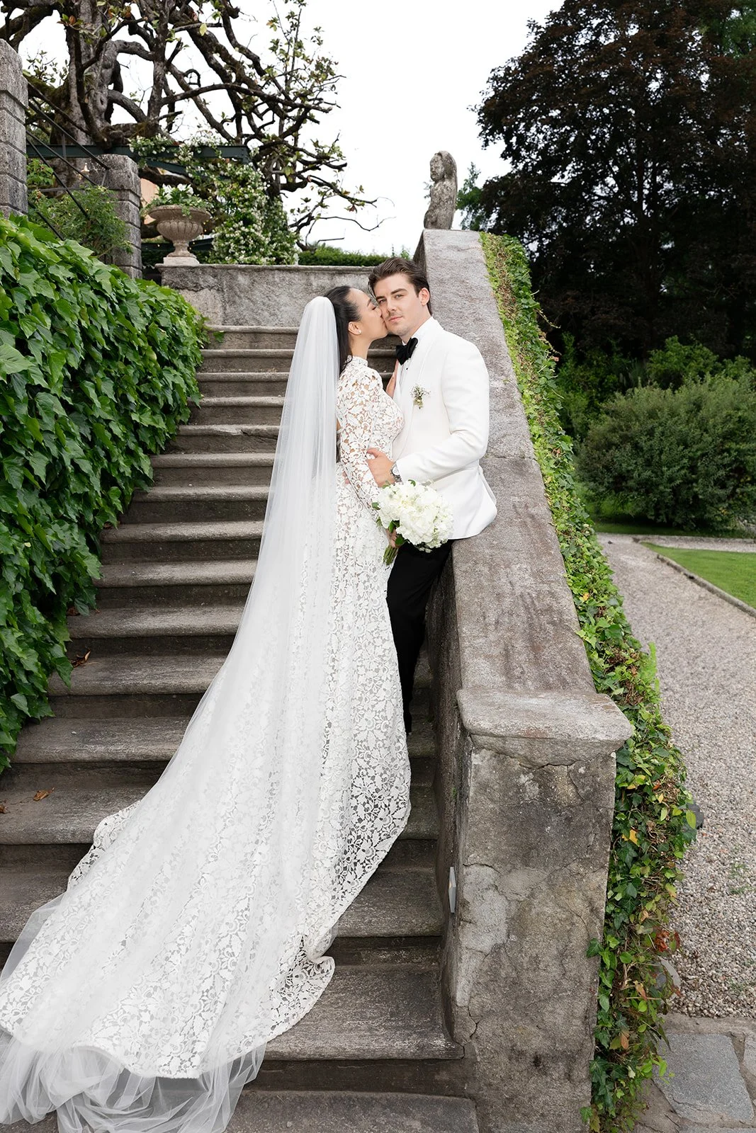 Ein Brautpaar posiert auf einer Steintreppe in einem Garten, die Braut trägt ein langes, weißes Spitzenkleid, der Bräutigam trägt einen weißen Anzug mit schwarzer Fliege, er hält einen Blumenstrauß.
