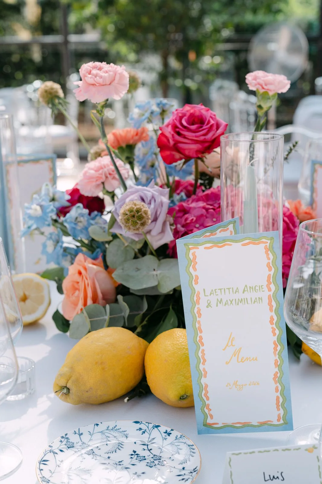 Blumenstrauß mit rosa, lila und blauen Blumen, Zitronen auf einem festlich gedeckten Tisch bei einer Hochzeit im Freien, mit Namensschild für Laetitia Angie & Maximilian, Datum 24. Mai 2023