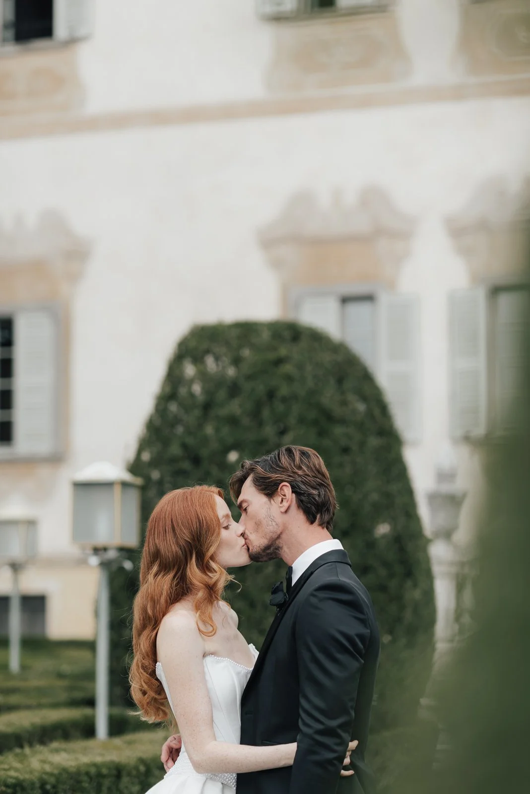 Ein Paar in formeller Kleidung, wahrscheinlich bei einer Hochzeit, küsst sich vor einem großen, grünen Busch und einem weißen Gebäude mit Fensterläden.