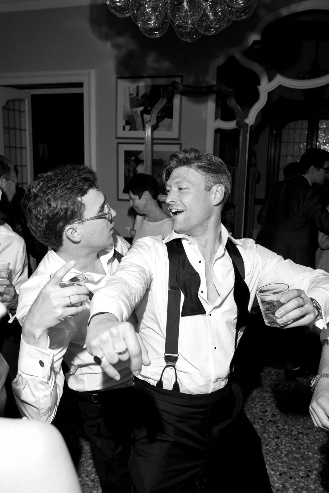 Zwei Männer lachen und feiern bei einer Party, einer trägt ein weißes Hemd und hat eine Kamera um den Hals, der andere trägt Brille und hält ein Glas in der Hand.