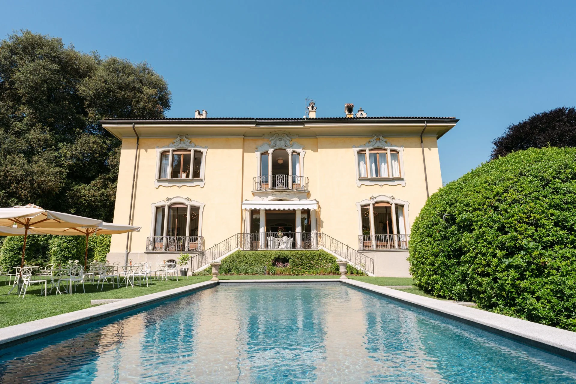 Elegantes, gelbes Herrenhaus mit Terrassen, Balkon und großen Fenstern, umgeben von grünem Garten und Pool, unter klarem blauen Himmel.