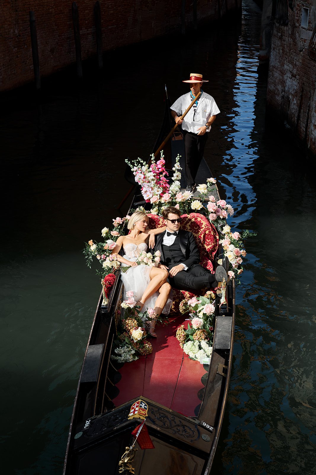 Ein Paar in Hochzeitskleidung auf einer mit Blumen geschmückten Gondel in Venedig, gesteuert von einem Gondoliere in weißem Hemd und Strohhut.