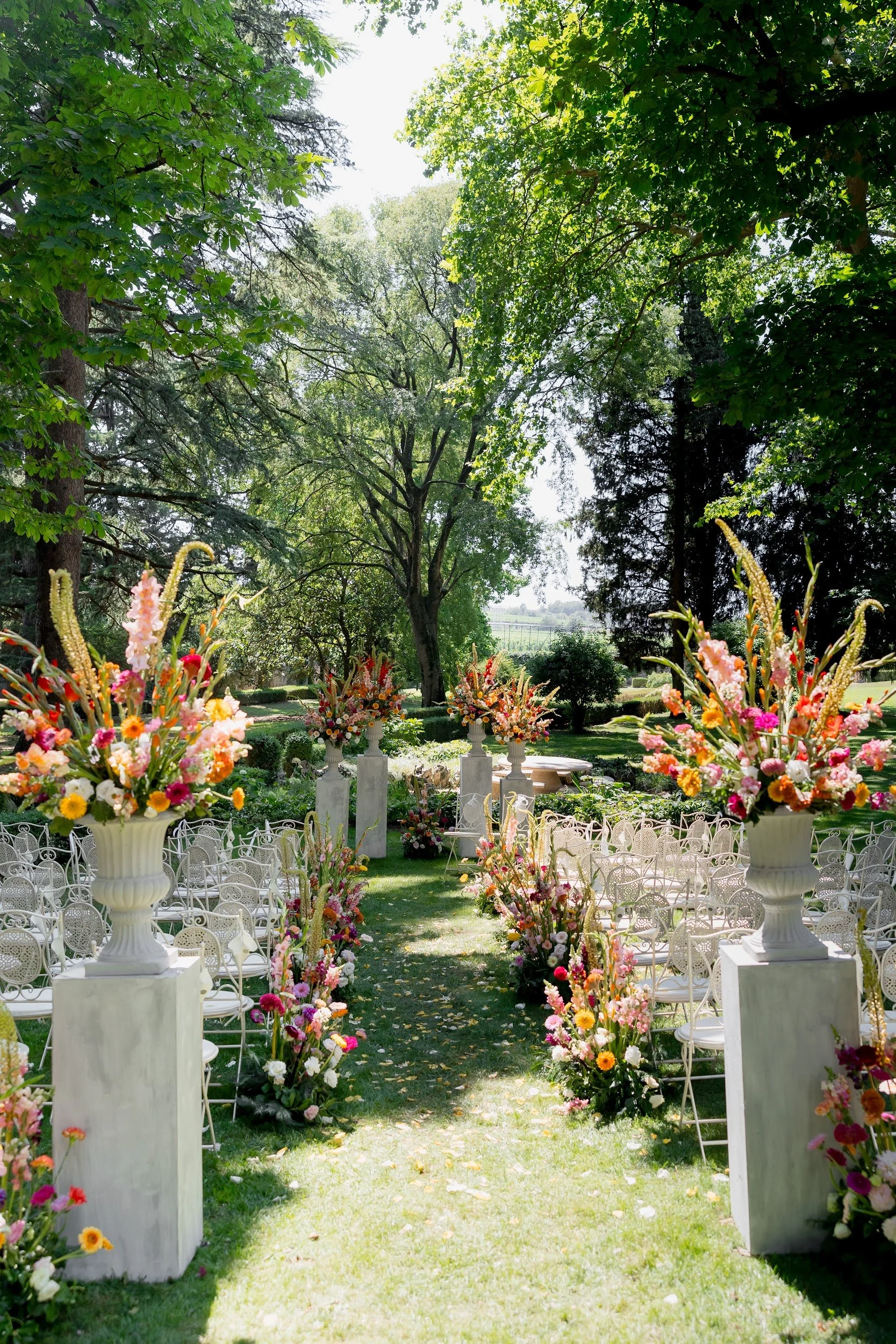 Ein Garten ist für eine Hochzeitszeremonie dekoriert, mit weißen Stühlen, großen Blumenvase mit bunten Blumen, und einer Allee aus Blumen und Statuen, umgeben von grünen Bäumen, bei Sonnenlicht.