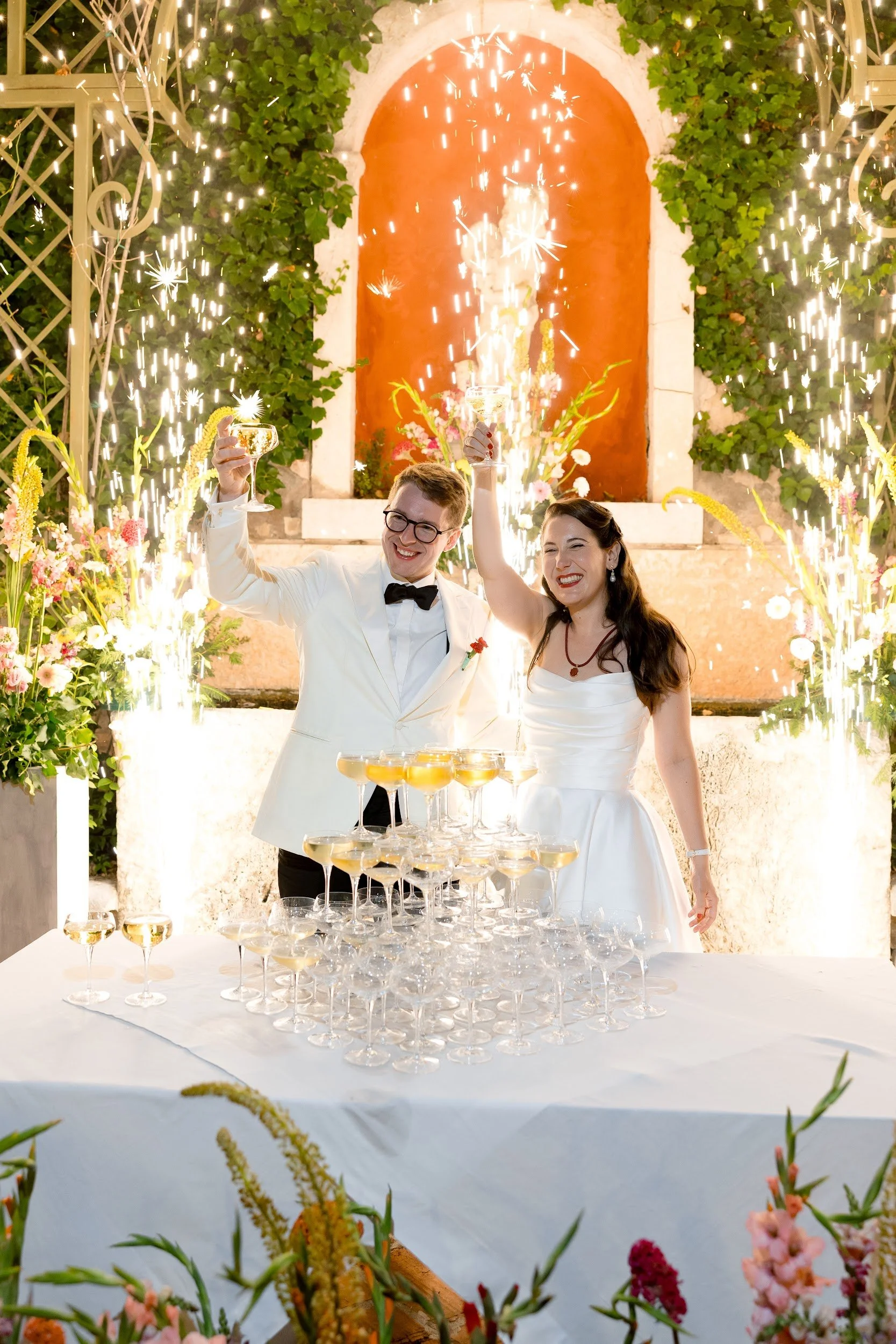 Ein frisch verheiratetes Paar bei seiner Hochzeitsfeier, das mit Sekt anstößt, vor einer Fountain- und Blumen-Dekoration, mit Sektgläsern auf einem gedeckten Tisch und Feuerwerkskörpern im Hintergrund bei Nacht.