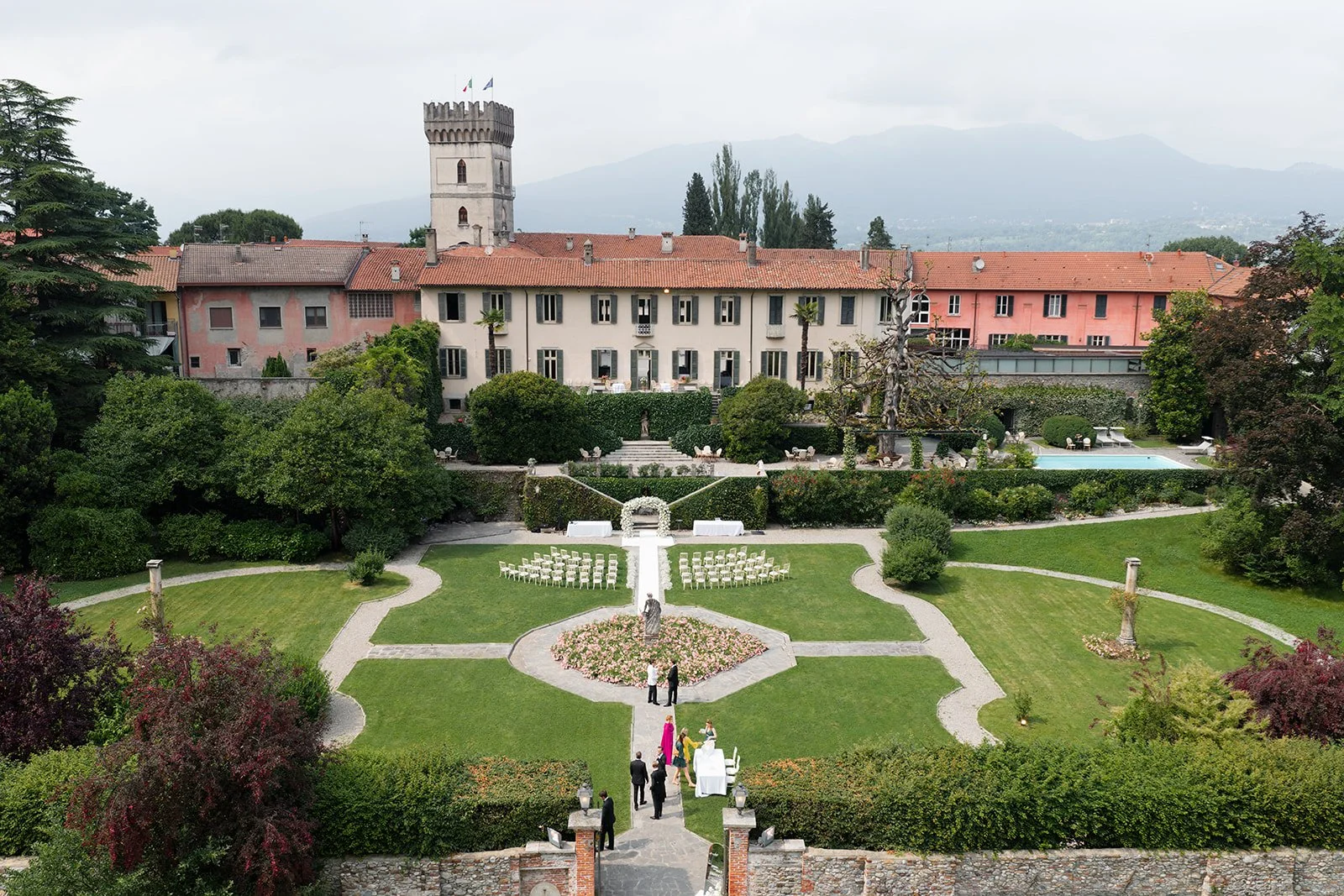 Garten mit zeremoniellem Sitzbereich, Blumen, Statuen und einer hölzernen Pergola, im Hintergrund ein altes Gebäude mit Turm