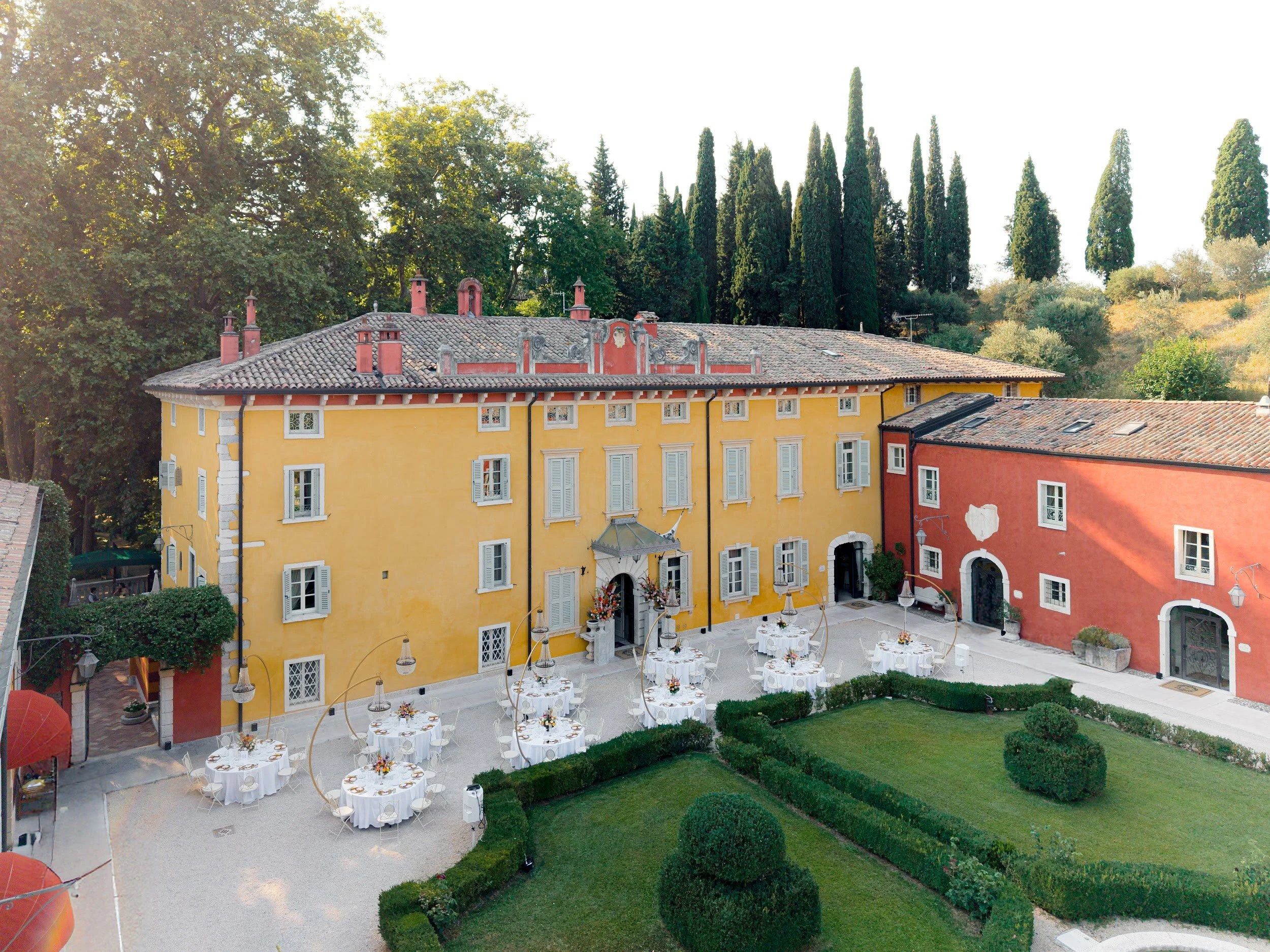 Ein gelbes Gebäude mit roten Akzenten und mehreren Fenstern. Im Vordergrund sind weiß gedeckte Tische mit Blumen, sowie ein gepflegter Garten mit Büschen und Rasen. Im Hintergrund sind hohe, schlanke Bäume und ein blauer Himmel.