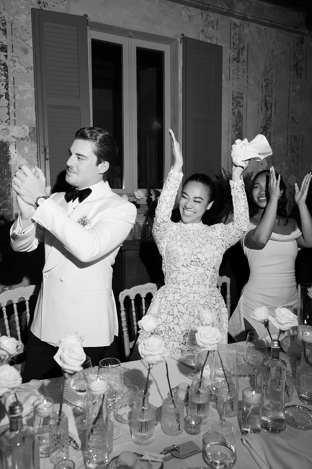 Menschen feiern eine Hochzeit, einer lächelt mit verschränkten Händen, die anderen lachen und klatschen, im Hintergrund eine festliche Tischdekoration mit Blumen und Kerzen.