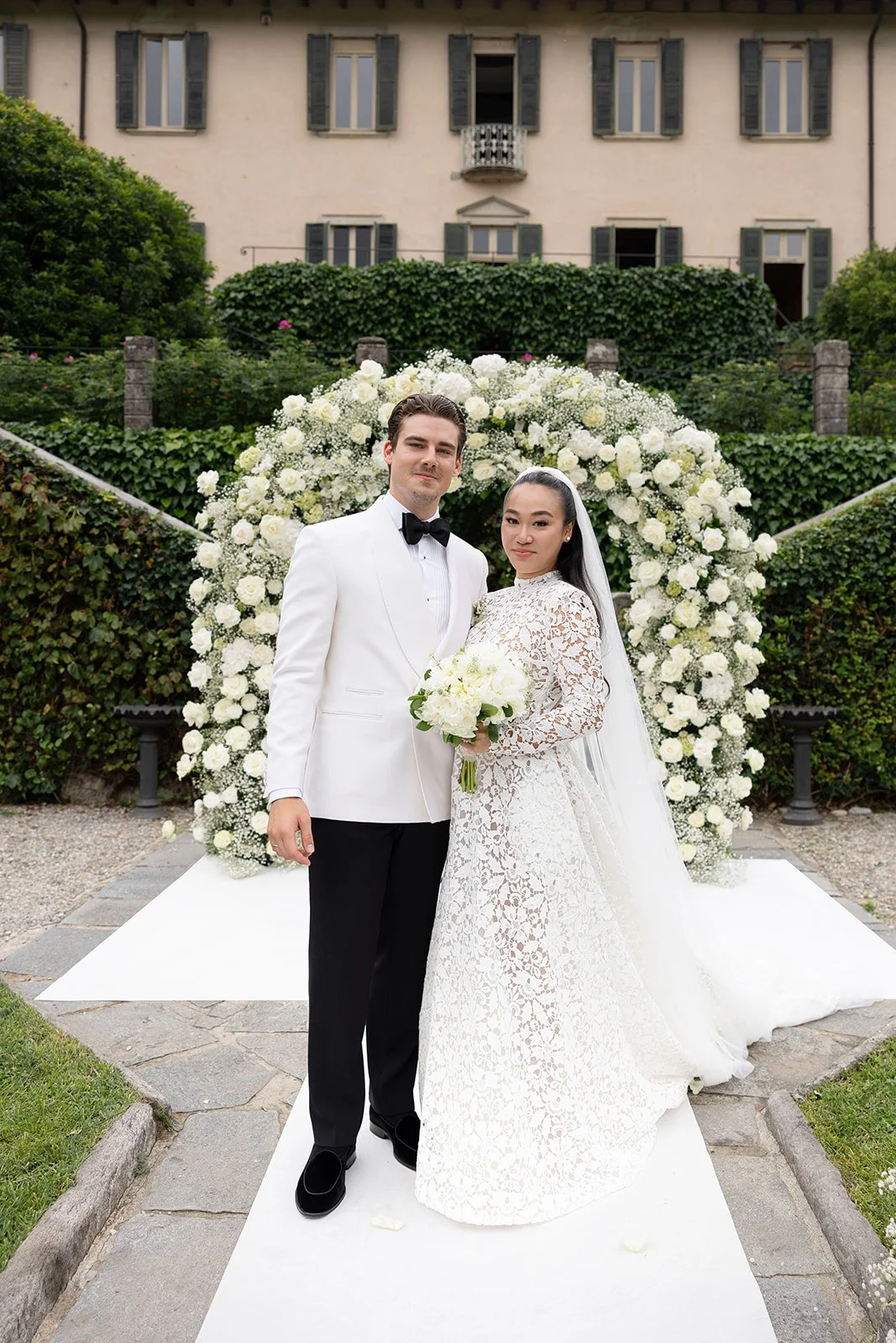 Ein Brautpaar bei einer Hochzeit, stehend vor einem Blumenbogen aus weißen Blumen, im Garten eines Hauses mit mehreren Fenstern und grünen Fensternläden.