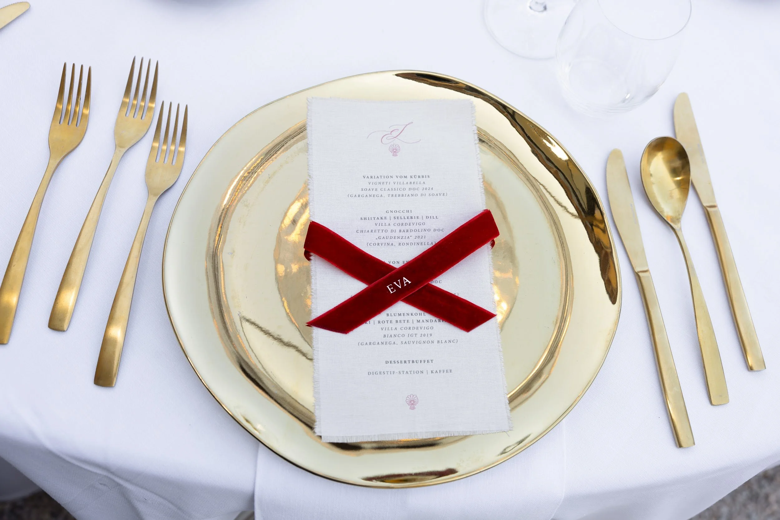 Ein eleganter gedeckter Tisch mit goldfarbenem Besteck, einem goldfarbenen Teller und einem weißen Servietten mit einem Menüblatt, das mit rotem Band mit dem Namen 'EVA' verziert ist.