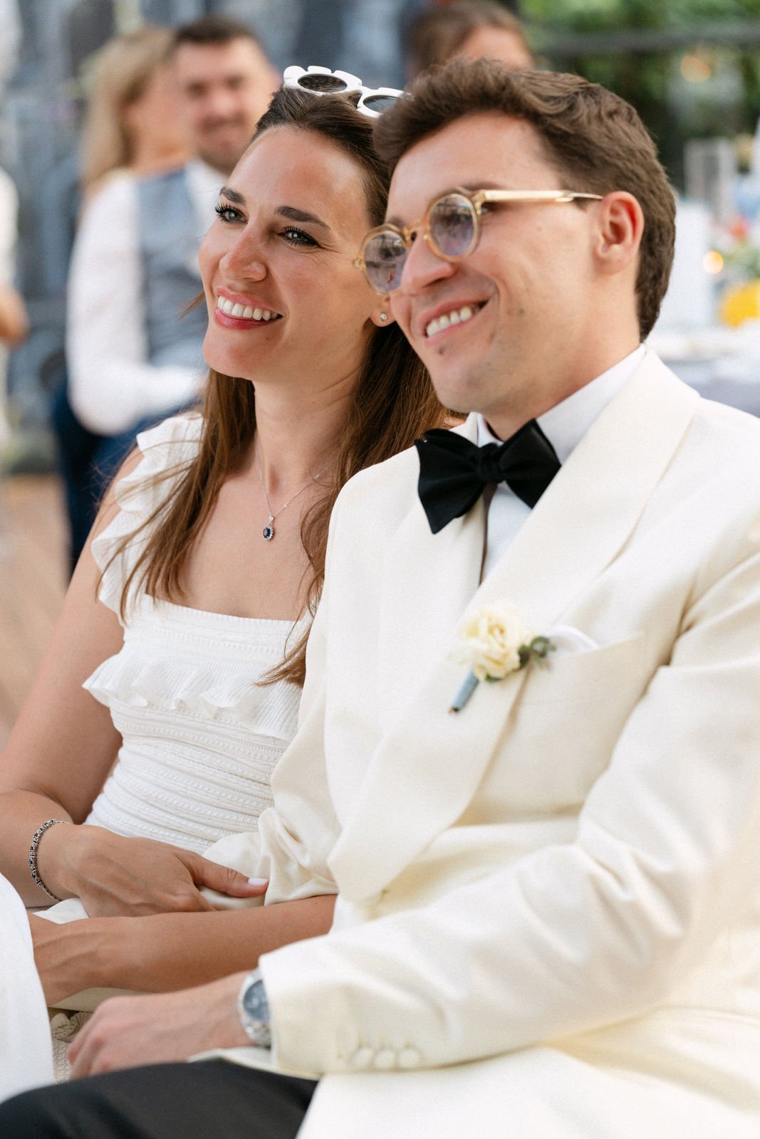 Ein glückliches Brautpaar sitzt bei einer Hochzeit, die Frau trägt ein weißes Kleid und eine Halskette, der Mann trägt einen weißen Anzug mit Fliege und Brille.