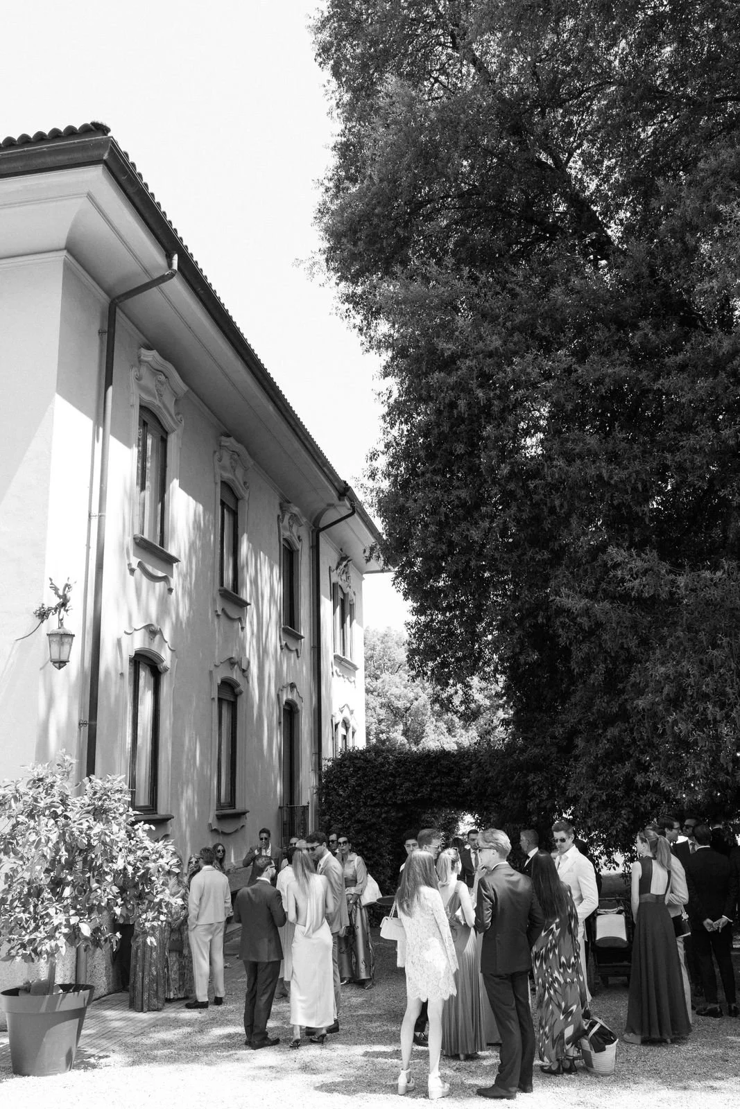 Gruppe von Menschen im Freien vor einem großen Haus, einige stehen und sprechen, bei schönem Wetter, Schatten von Bäumen