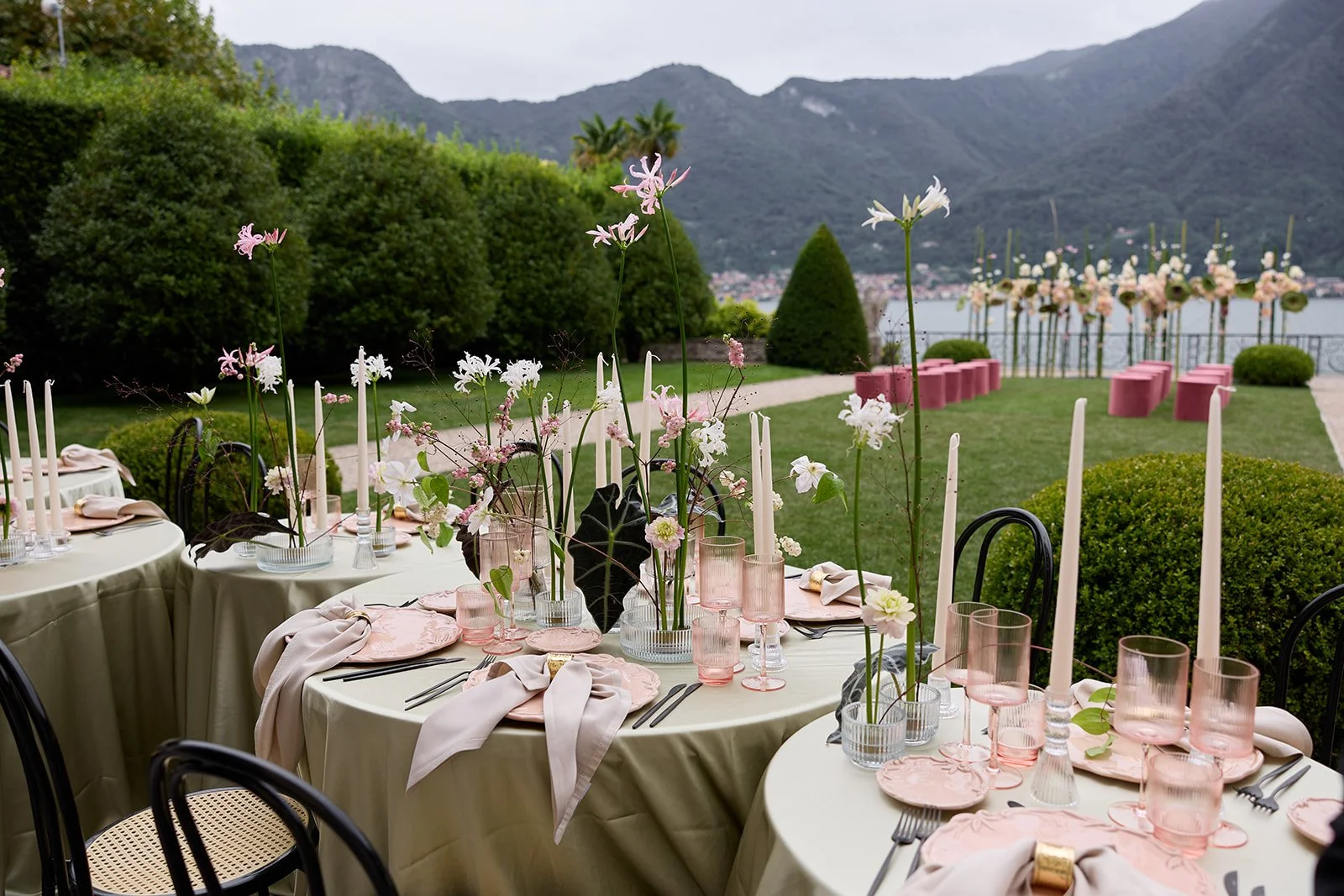 Elegante Außenveranstaltung mit runden Tischen, rosa und weißen Dekorationen, Blumentischarrangements und Blick auf einen Garten und Berge im Hintergrund.