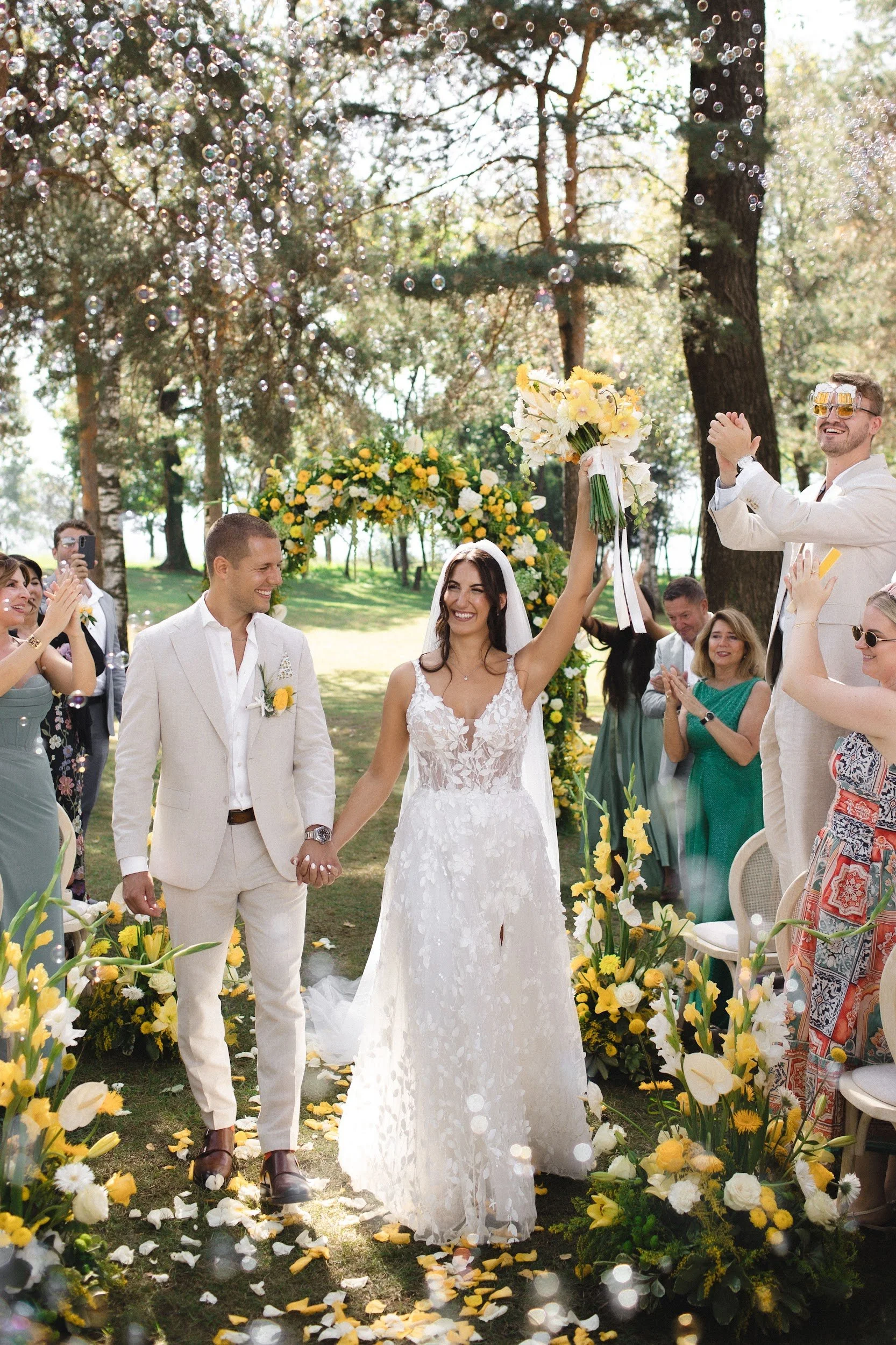 Ein glückliches Brautpaar beim Hochzeitszeremoniestil in einem Wald, umgeben von Familienmitgliedern und Freunden, mit Blumen und Luftballons, die bei Sonnenschein auf einem Rasen unter Bäumen feiern.