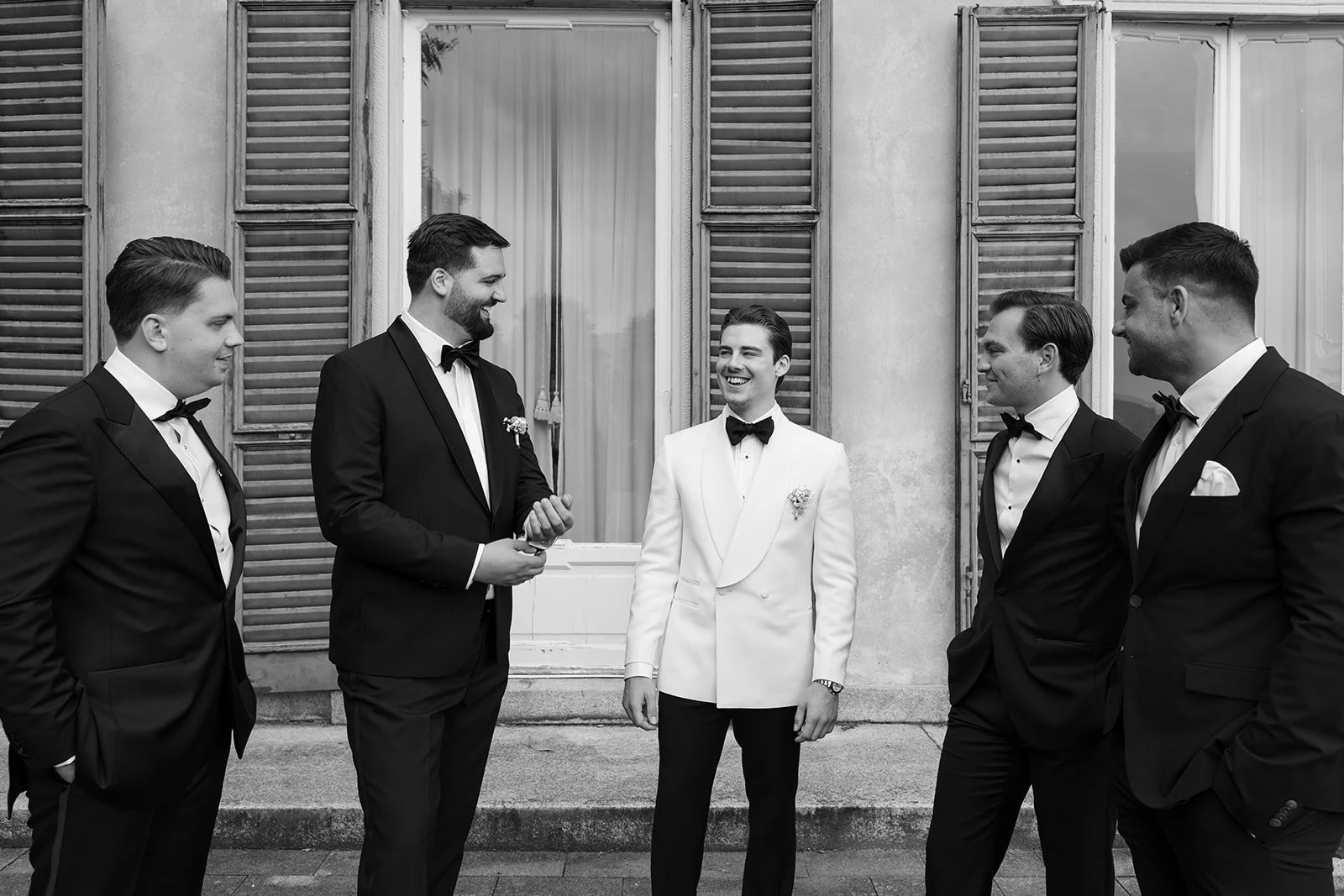 Männer in formeller Kleidung, vermutlich bei einer Hochzeit, stehen vor einer alten Gebäudefassade mit offenen Fensterläden, im Gespräch und lachen.