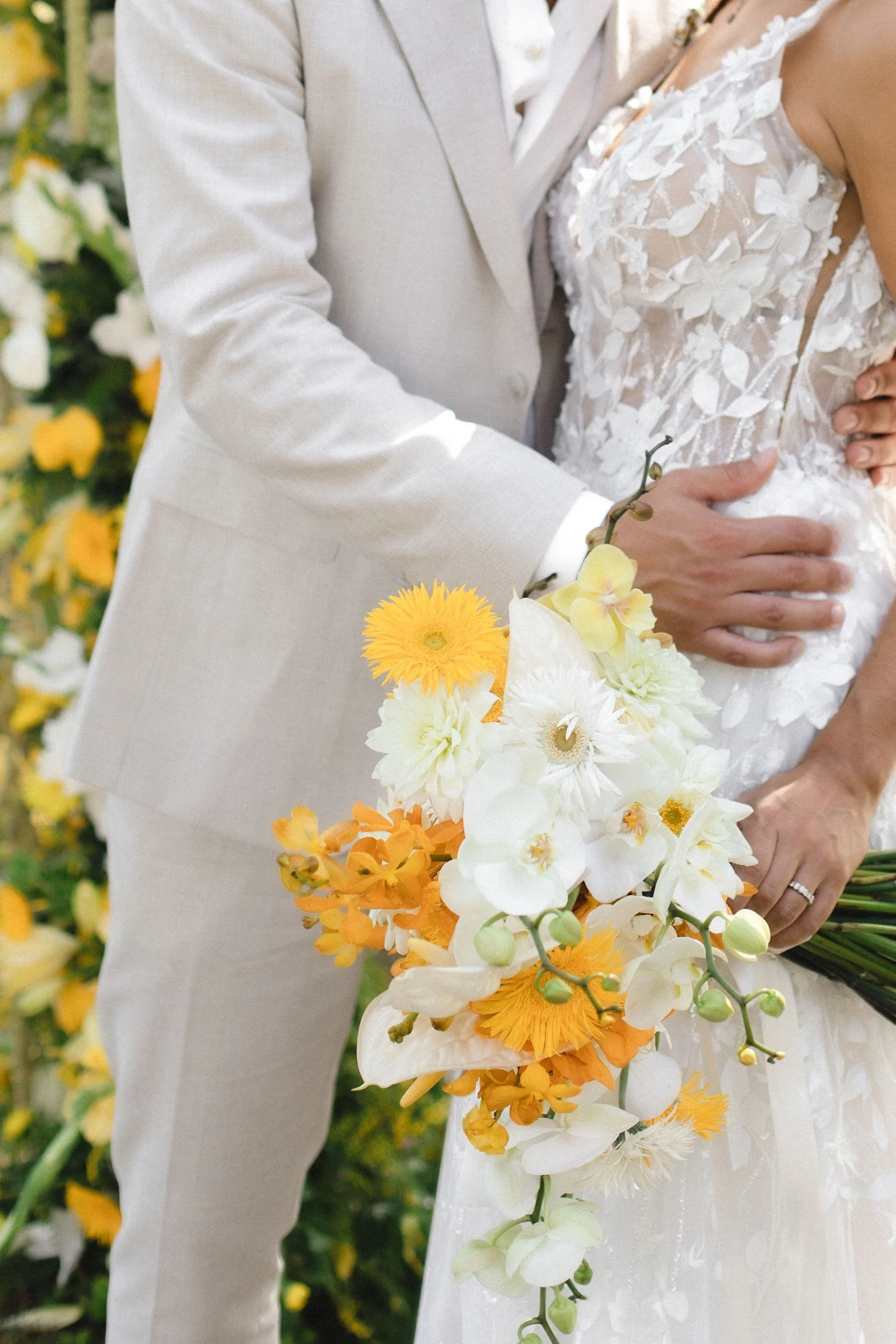 Nahaufnahme eines Hochzeitspaars, das sich umarmt, mit Fokus auf ihre Kleidung und das Brautstrauß, vor einer Kulisse voller gelber und weißer Blumen.