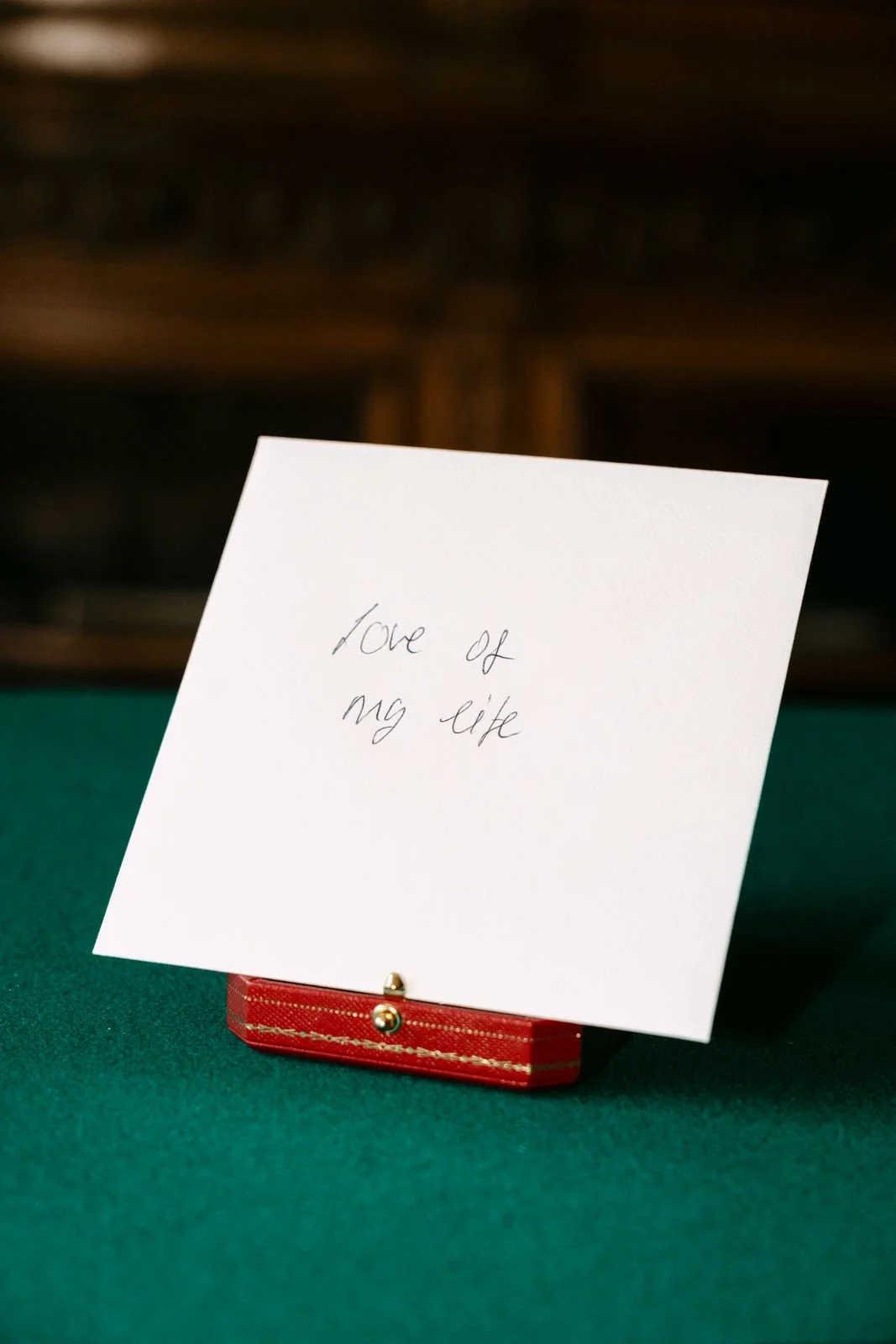 Ein weißer Zettel mit dem handgeschriebenen Text 'love of my life', der auf einem roten Etui auf einem grünen Tischtuch steht.
