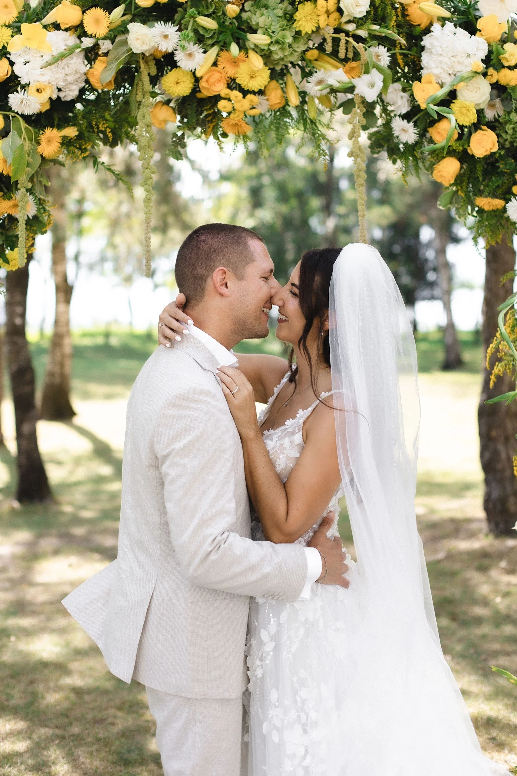 Ein Brautpaar bei einer Hochzeit, umgeben von einem Blumenbogen aus gelben, weißen und cremefarbenen Blumen, im Freien mit Bäumen im Hintergrund.