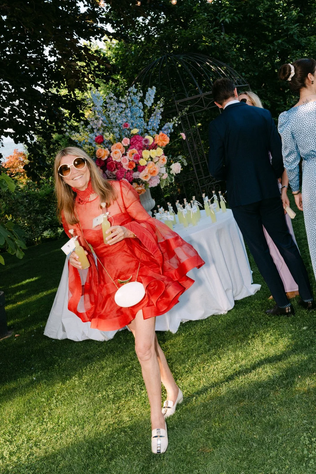Eine lachende Frau in einem roten Kleid mit großen Sonnenbrillen hält zwei Flaschen mit Getränken und posiert vor einem festlich gedeckten Tisch mit bunten Blumen, im Garten bei Sonnenlicht.