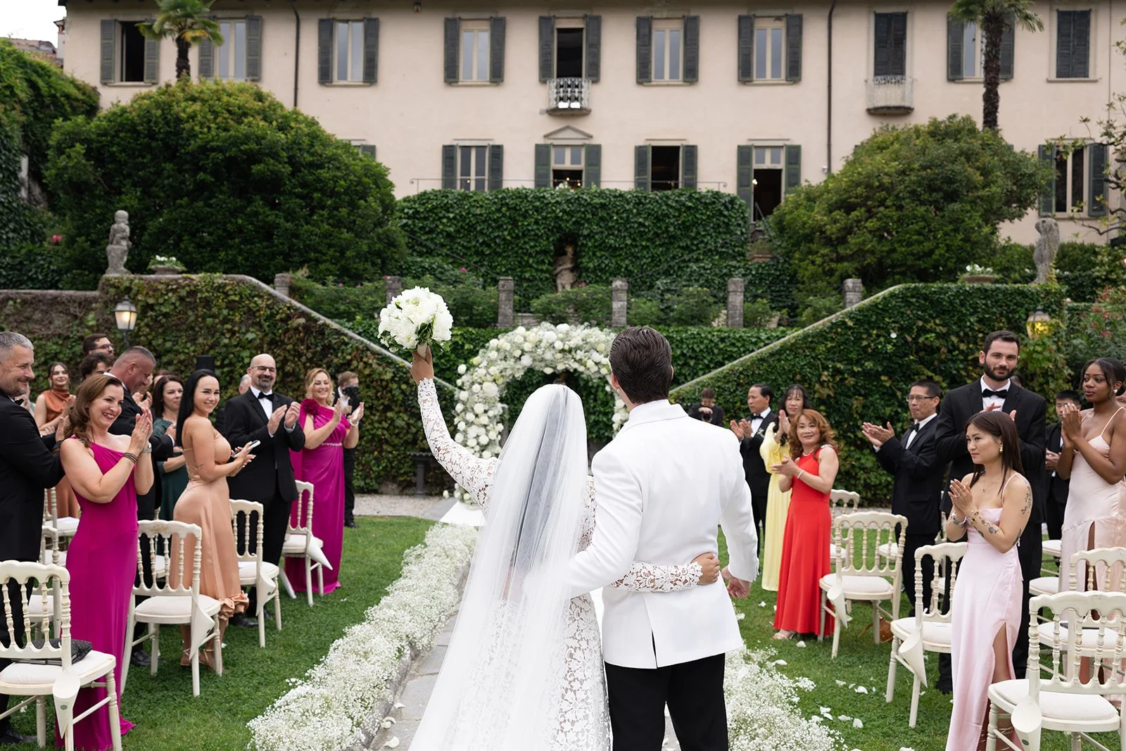 Ein Brautpaar bei ihrer Hochzeit im Garten, umgeben von Freunden und Familie, mit einem blühenden Landschaftshintergrund.