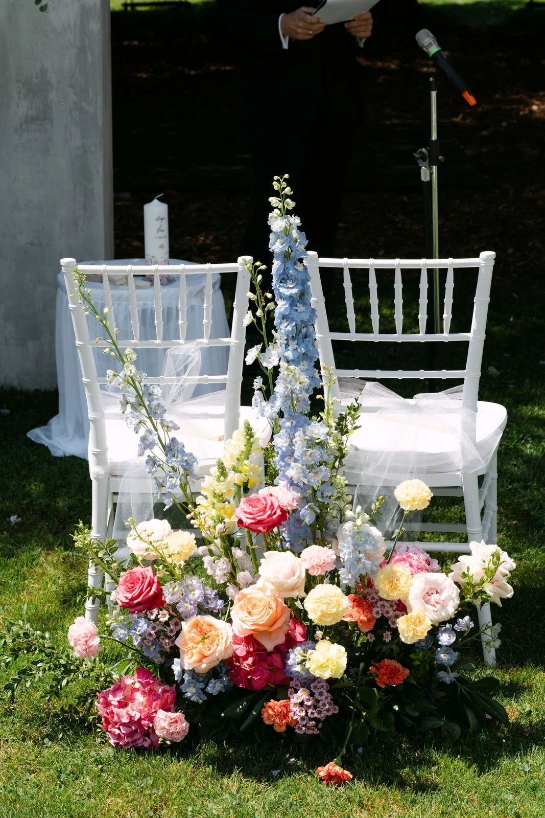 Ein weißes Blumenarrangement mit Rosen, Gladiolen und anderen Blumen vor weißen Stühlen bei einer Hochzeitszeremonie im Freien.