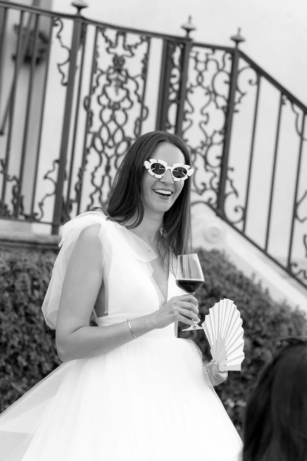 Lächelnde Frau im Hochzeitskleid mit Sonnenbrille und Weinglas, hält Fächer, vor einem Metallzaun, schwarz-weiß Foto.