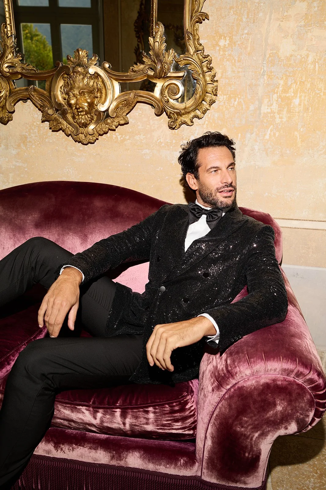 Ein Mann in formeller Kleidung sitzt auf einem pinkfarbenen Samtsofa vor einem goldfarbenen, barocken Spiegel mit Löwenkopf-Relieft. Die Wand im Hintergrund ist cremefarben mit Textur.