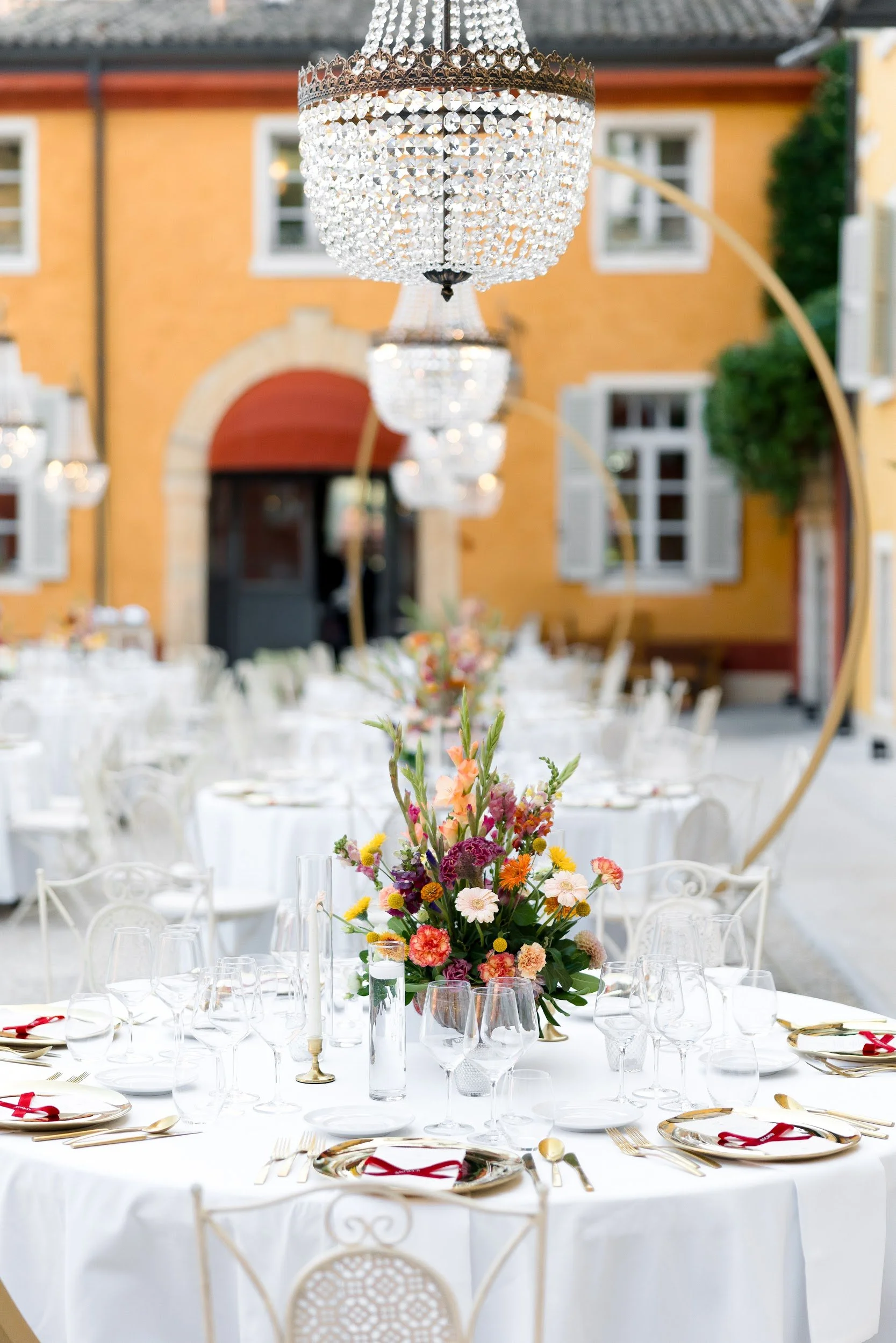 Festlich gedeckter Tisch mit buntem Blumenarrangement in der Mitte, Kerzen, Weingläser und goldene Bestecke in einem eleganten Außenbereich mit Blick auf ein gelbes Haus.