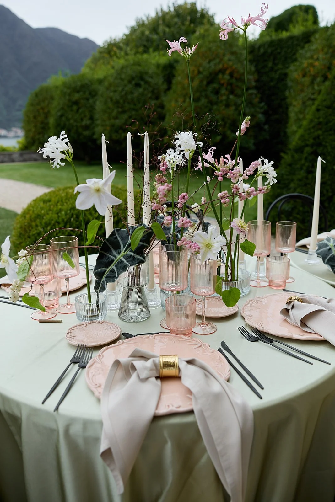 Eleganter gedeckter Tisch mit rosa Tellern, weißen Servietten, schwarzen Besteck, rosa Trinkgläser und eine Blumenmitte mit weißen und pinken Blumen, umgeben von Kerzen auf einer Wiese mit Bäumen und Bergen im Hintergrund.