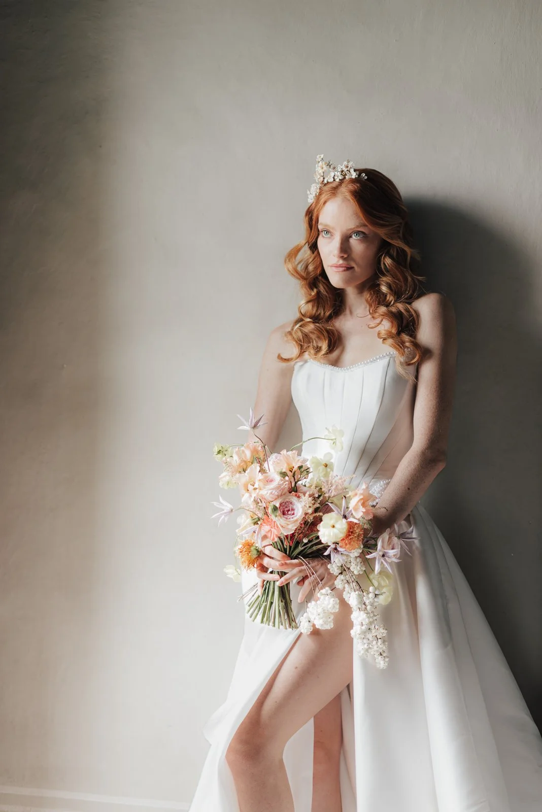 Eine rothaarige Frau in einem weißen Brautkleid mit Blumenkrone, die einen Blumenstrauß hält und an einer grauen Wand steht.
