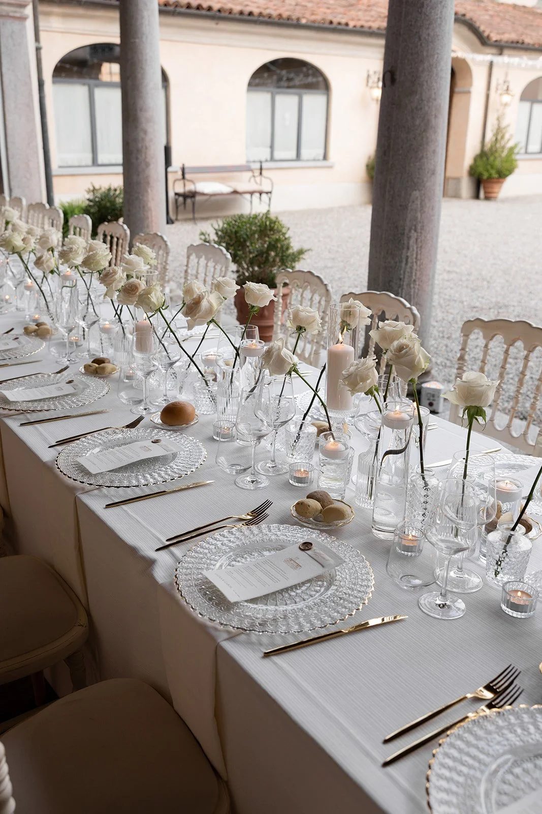 Elegant gedeckter Esstisch mit weißen Rosen, Kerzen, Gläsern, Tellern und Goldbesteck in einer offenen Veranda mit Blick auf ein Gebäude im Hintergrund.