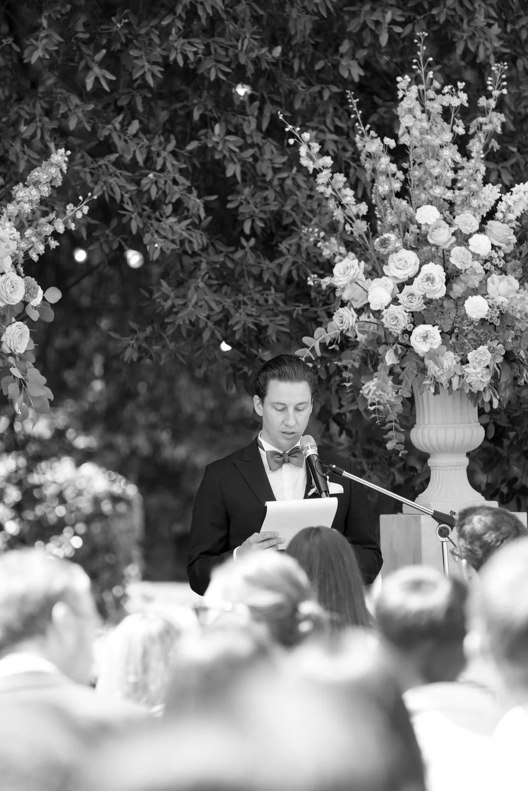 Ein Mann in formeller Kleidung, der an einem Redepult liest, bei einer Veranstaltung im Freien mit Blumentopf im Hintergrund und einem Publikum im Vordergrund.