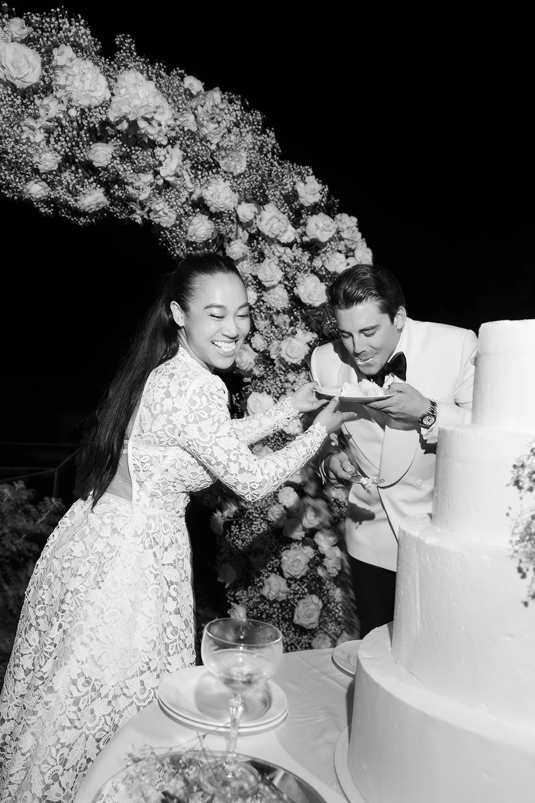 Ein Brautpaar bei ihrer Hochzeit, die Braut erhält ein Stück Hochzeitstorte vom Bräutigam, im Hintergrund eine Blumenüberraschung.